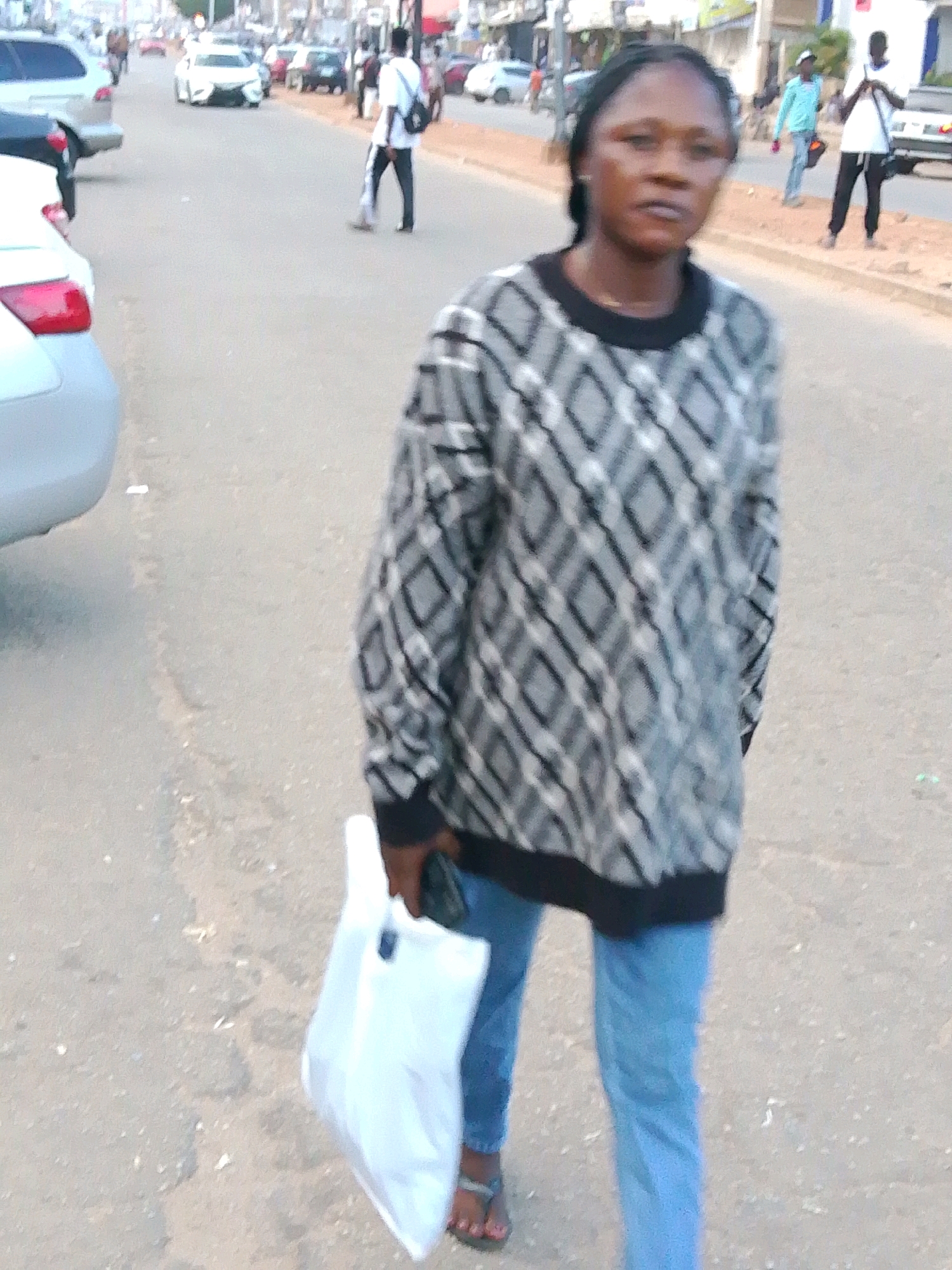 A woman, who appears to be in her late 20s or early 30s, walks on a paved street in Jos, Nigeria. She is wearing a grey and black patterned sweater and blue jeans, carrying a white plastic bag in her left hand. The street is lined with vehicles and pedestrians, suggesting it is a busy urban area. The woman is walking towards the camera, and her face is visible. She has dark hair and is looking forward. The background shows a typical street scene in Nigeria, with buildings and trees visible. The image seems to have been taken during the daytime.