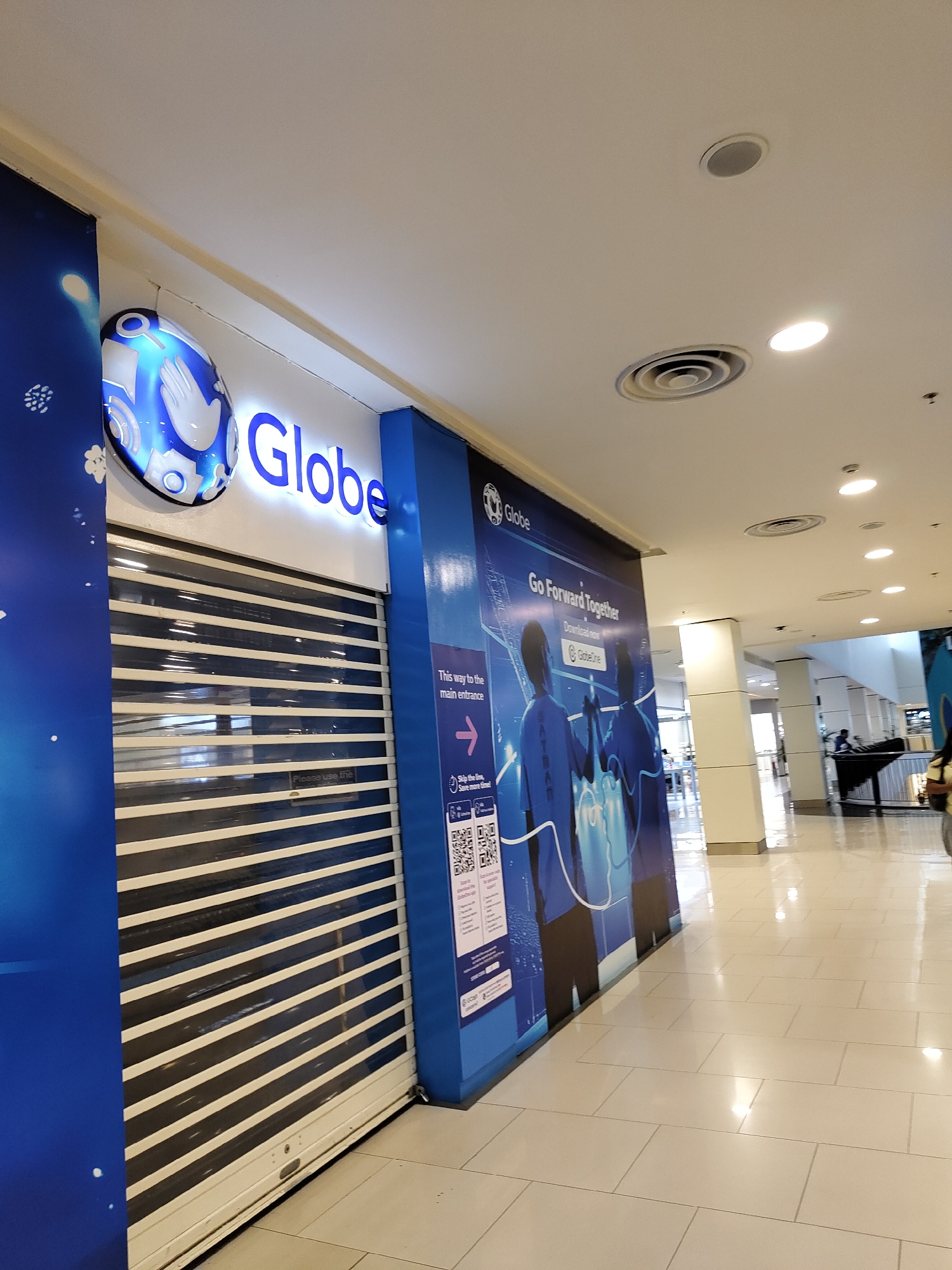 The image shows the exterior of a closed Globe store, a telecommunications company, in a mall in Davao City, Philippines. The store is identifiable by its blue branding and signage, including the "Globe" logo and the slogan "Go Forward Together." A large banner on the right side of the store displays images of people and promotional text. The store's entrance is covered by a metal roll-up shutter, indicating it is not open for business. The floor is tiled and appears to be a common area within a mall, with a light-colored, reflective surface. In the background, other parts of the mall are visible, including columns, ceiling lights, and air vents. There are no people directly interacting with the Globe store itself. The overall environment suggests an indoor shopping mall setting.