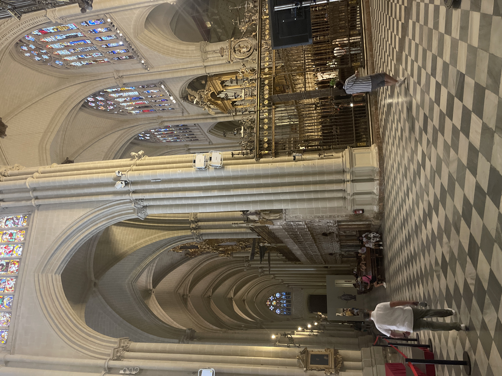 The image captures the interior of the Toledo Cathedral in Toledo, Spain. The vast nave features towering stone columns and ribbed vaults, characteristic of Gothic architecture. Stained-glass windows, brightly colored, are visible along the upper walls. The floor is a dramatic black and white checkered pattern.
In the foreground, a man wearing a white shirt and green pants walks away from the camera, carrying a water bottle. To the right, a person in a striped shirt and shorts walks away from the camera. In the middle ground, several people are seated on pews, facing a golden choir screen and an organ. The overall scene depicts a public space within the cathedral, with visitors observing the architecture and ornate interior features.