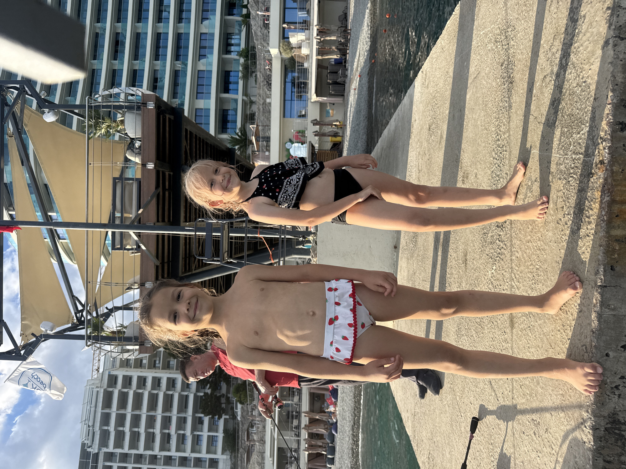 Two young girls are standing on a concrete walkway next to a body of water. The girl in the foreground is wearing white bikini bottoms with strawberries on them. The girl behind her is wearing a black bandana-style bikini top and black bottoms. Both girls are smiling. Behind them, a modern hotel building with many windows is visible. The sky is blue with some clouds. The sun is shining, casting shadows on the walkway. A flag with text on it is partially visible on the left. There is also a person in a red shirt with a fishing rod visible in the background. The scene appears to be taking place in a resort or coastal area, possibly Yalta, Ukraine, during the daytime.