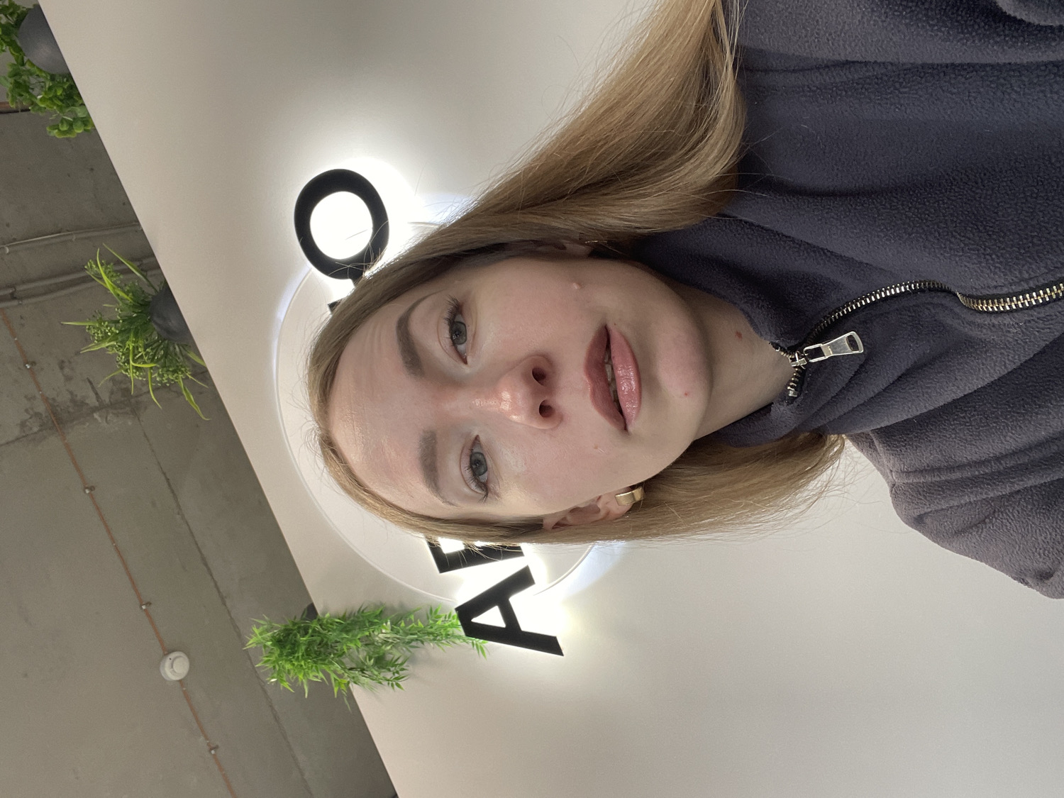 This indoor selfie, taken with a sideways orientation, features a young woman with fair skin, light-colored eyes, and shoulder-length blonde or light brown hair. She has a neutral expression, with lightly defined eyebrows and lips, which appear glossy. She is wearing a dark grey or dark blue fleece jacket with a silver zipper, partially unzipped. A small gold-colored earring is visible on her left ear.
The setting appears to be a commercial or public space in Perm, Russia. The background reveals a white ceiling surface with a large, illuminated circular element, possibly a light fixture or part of a logo. Adjacent to this, large black letters are visible on what seems to be a sign, including a clear "A" and another round shape that could be an "O" or part of a character, like the top of an "L".
Above the woman's head (due to the image's rotation), an exposed concrete ceiling with visible pipes or conduits and a white smoke detector can be seen. Two artificial green plants, hanging in dark pots, add a touch of greenery to the industrial-style ceiling. The overall lighting is bright and artificial.