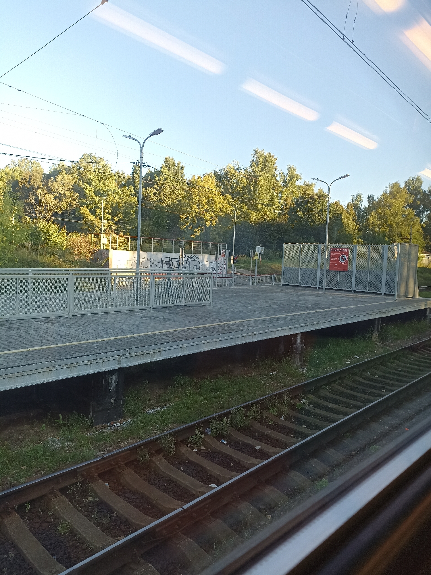 The image depicts a train station platform in Podolsk, Russia, on a clear day with a bright sky. The platform is constructed with gray paving stones and is bordered by a light-colored metal railing. A few streetlights are visible on the platform, and a tall fence encloses one section of the platform, displaying a red warning sign with text in Russian. Beyond the platform, a line of trees with green and yellow foliage is visible. Train tracks run in the foreground, with ballast and concrete sleepers. The scene is captured from the perspective of someone looking out of a moving train window, as indicated by the reflections in the glass.
