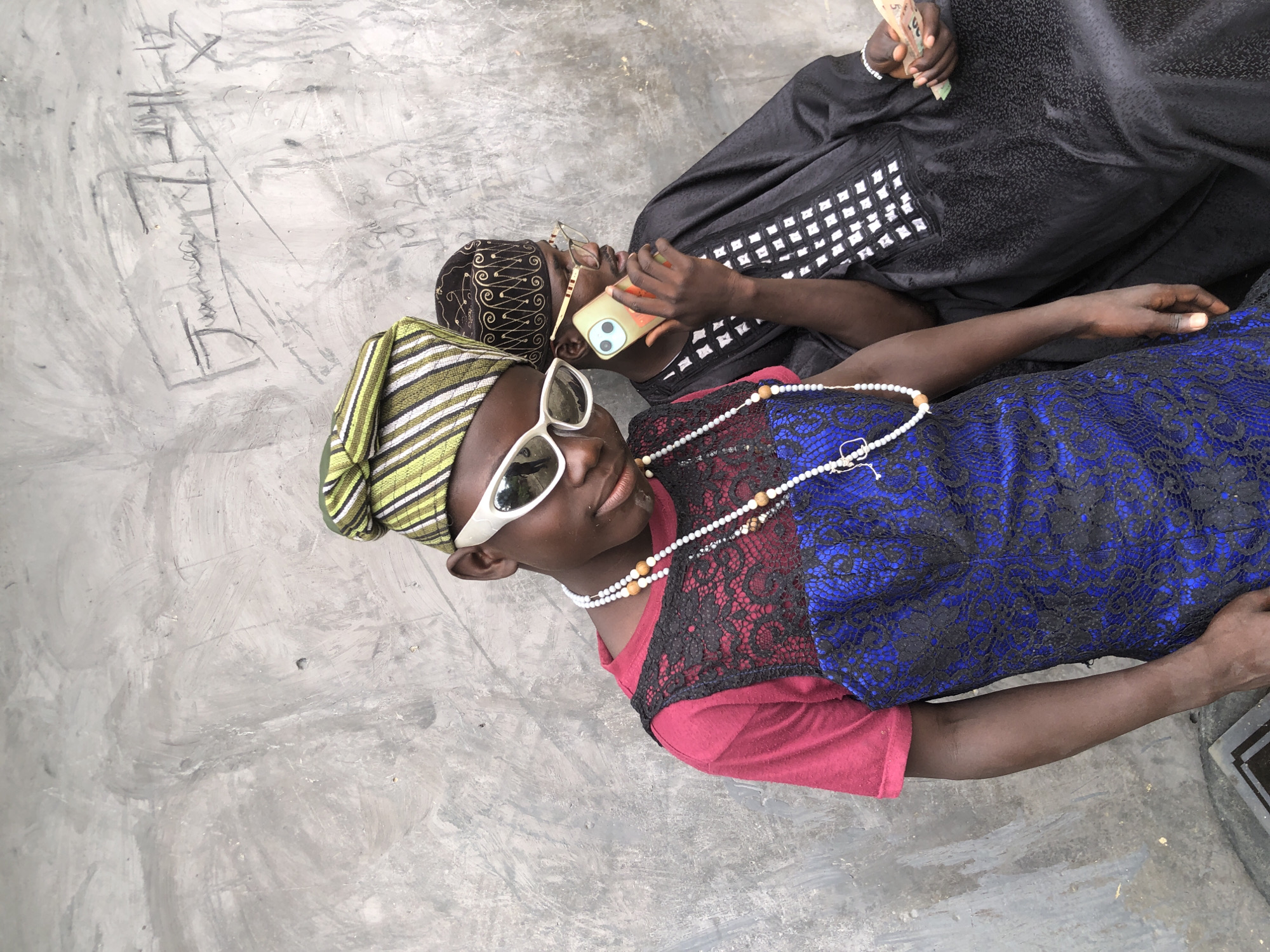 In Ado, Nigeria, two young men are pictured against a textured concrete wall with graffiti. The man in the foreground, on the left, wears a green and white striped cap, white-rimmed sunglasses, a maroon shirt, and a blue and black lacy dress over the shirt. He has a multi-strand beaded necklace with wooden and white beads. The man in the background, on the right, wears a dark cap with gold embroidery and a black robe adorned with white checkered patterns on the chest. He is holding a light-colored smartphone to his ear with his right hand and cash in his left hand. The overall lighting suggests an outdoor environment, possibly during daylight.