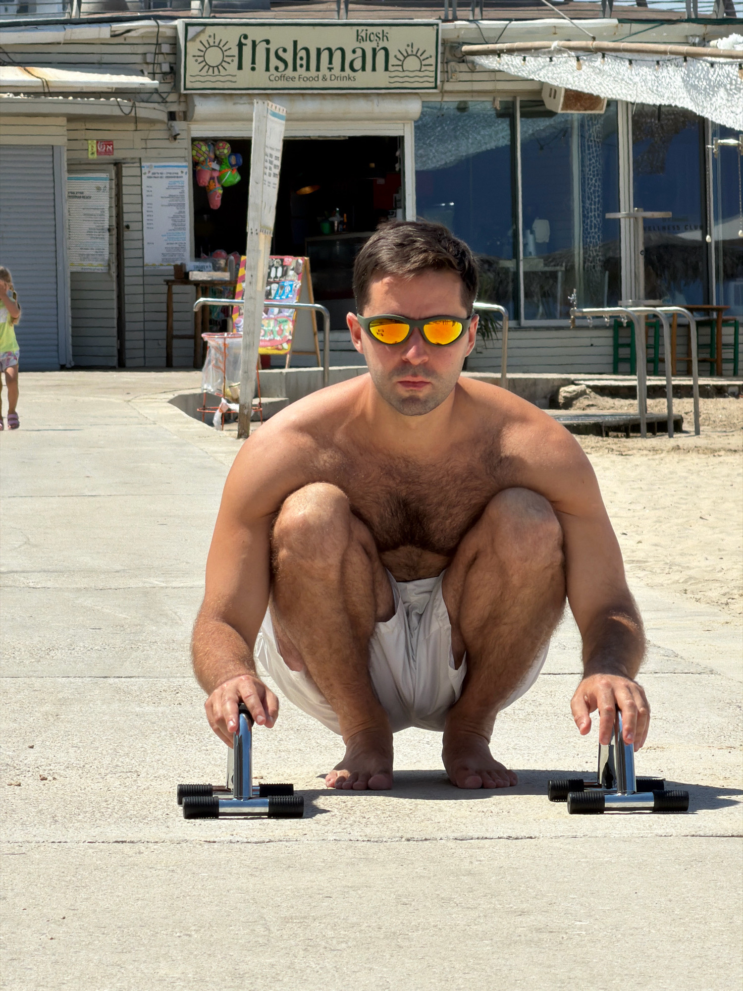 A man with dark hair and a light beard, wearing orange-tinted sunglasses and light-colored shorts, is crouched on a paved walkway with two metal push-up bars in front of him. His hands are positioned to grip the bars. His chest and abdomen are bare and show significant hair. In the background, a building with a sign reading "fishman Coffee Food & Drinks" is visible. To the left, a young girl in a yellow and pink outfit walks away from the camera. The scene appears to be a beachside area in Tel-Aviv, Israel, given the architecture and the presence of a walkway and sandy area.