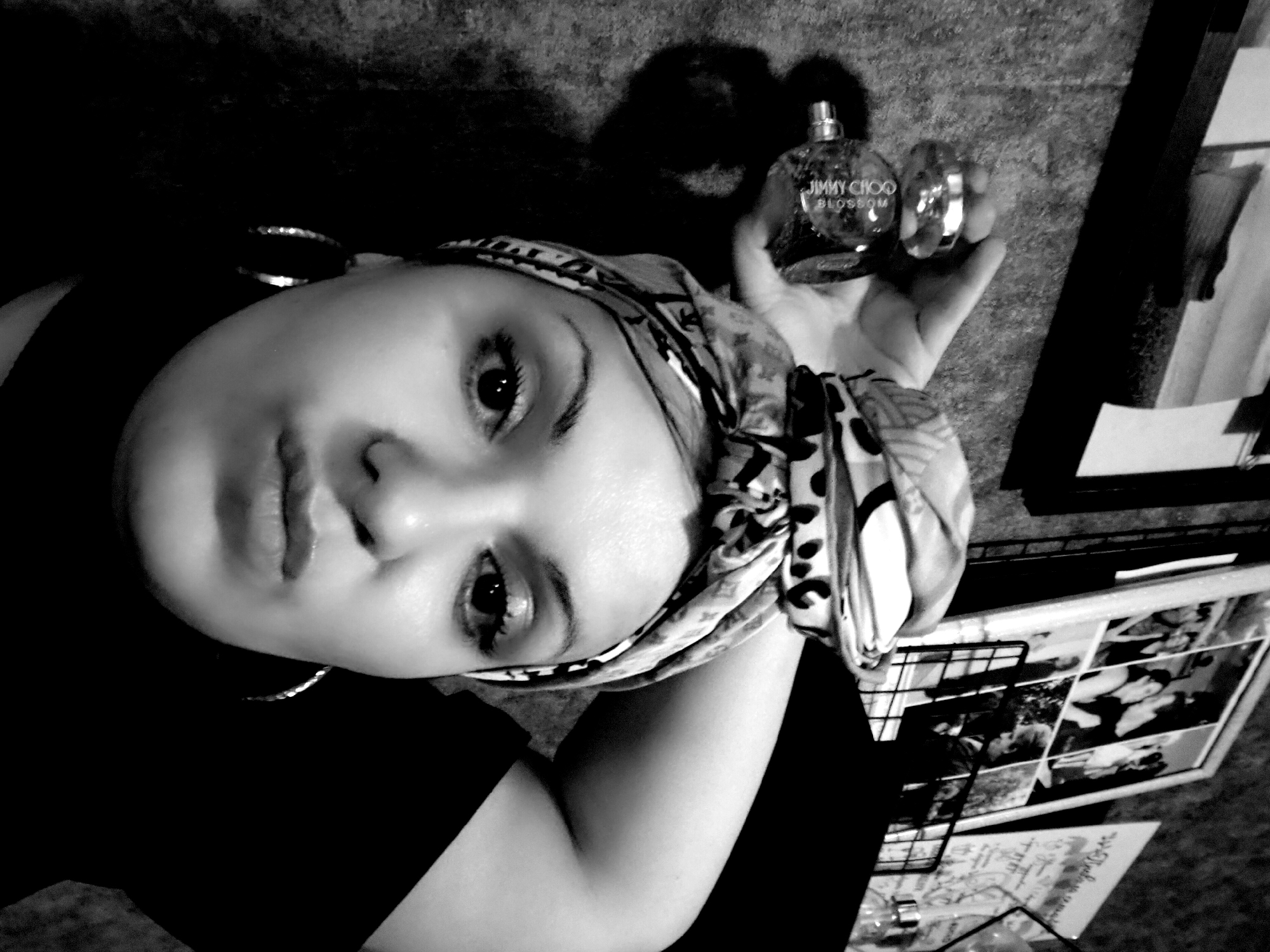 A black and white, medium-close-up photograph depicts a young woman with fair skin, dark eyes, and full lips looking directly at the camera. She is wearing a patterned headscarf and large hoop earrings. She holds a bottle of Jimmy Choo Blossom perfume in her right hand, near her head. The woman is wearing a dark-colored top. In the background, there is a wall and a framed photo collage. The location is Марковский, Russia.