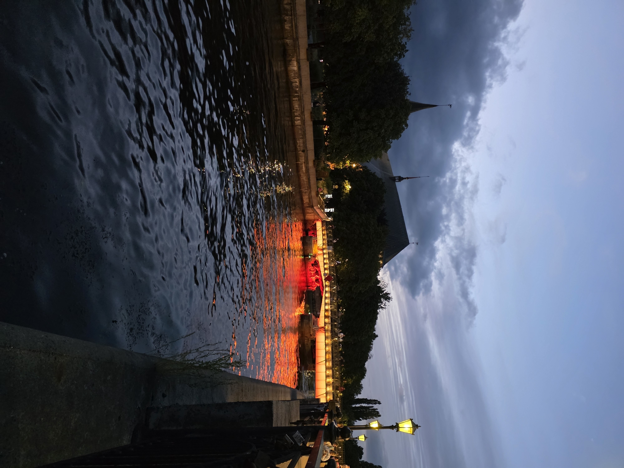 The image depicts a twilight scene over the Pregolya River in Kaliningrad, Russia. A bridge illuminated with red and orange lights spans the river, reflecting vibrantly on the water's surface. The bridge is lined with people, suggesting it is a pedestrian walkway or thoroughfare. On the left bank, a large, dark building with a pointed spire, likely a cathedral, is visible behind a line of trees. Traditional yellow streetlights are present along the right bank, casting a warm glow. The sky is a gradient of deep blue and grey, with scattered clouds adding texture. The overall atmosphere is serene and picturesque, capturing the beauty of the city at dusk.
