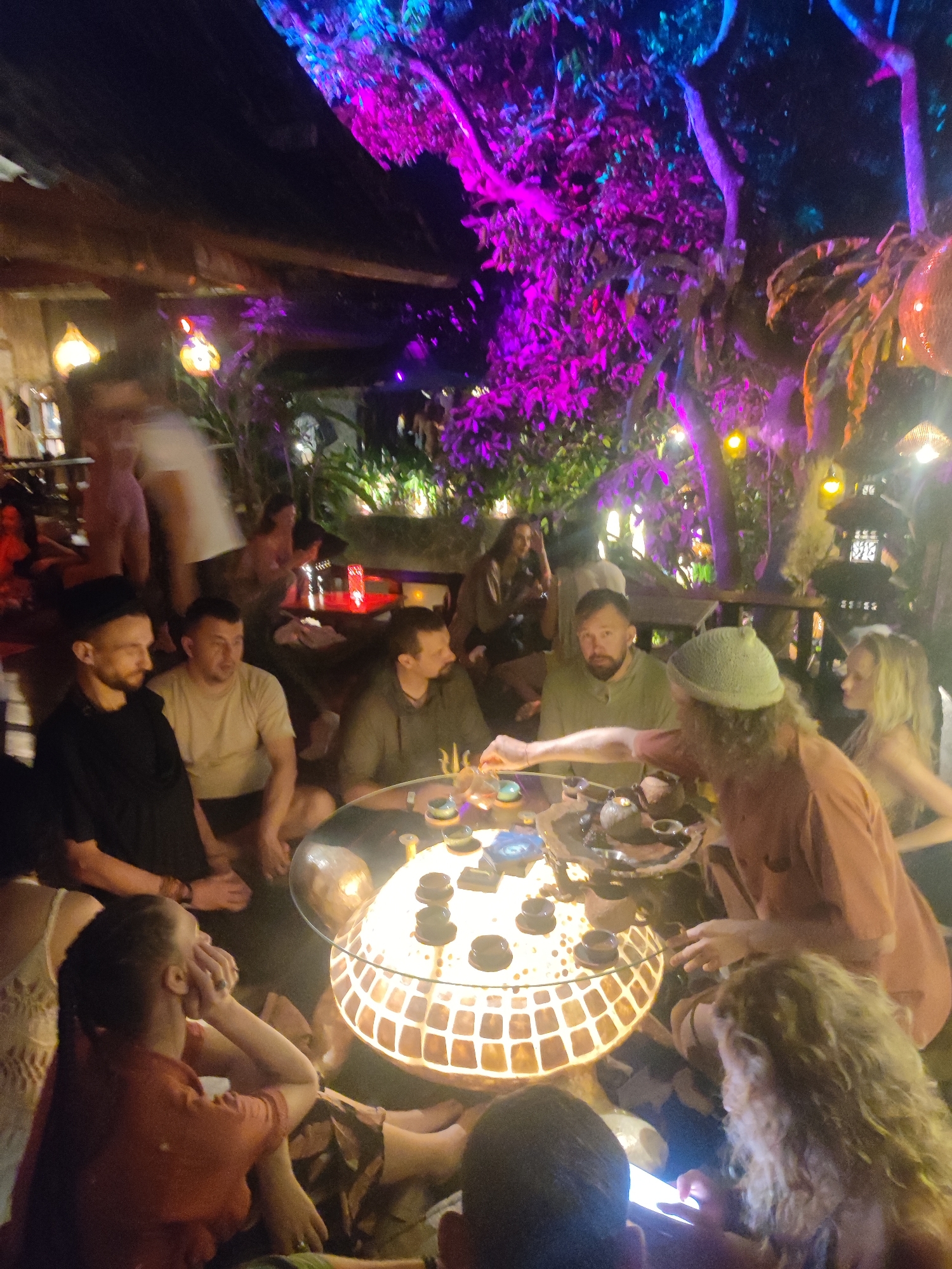 A group of approximately 15 people are gathered around a low, illuminated, round table for a tea ceremony in an outdoor setting, likely a cafe or private yard. The location is Lokaserena, Indonesia, judging by the lantern design and overall tropical atmosphere. The table is set with multiple small teacups, a teapot, and implements for preparing tea. One man, wearing a green beanie and an orange shirt, seems to be leading the ceremony, using a long, thin tool. The other participants are sitting on the ground, some observing the ceremony and others socializing. A woman with curly hair in the foreground is looking at her phone. The background is filled with lush vegetation, brightly lit with pink and blue lights, and decorative lanterns. A building structure with wood beams is partially visible in the background. The atmosphere appears relaxed and informal.