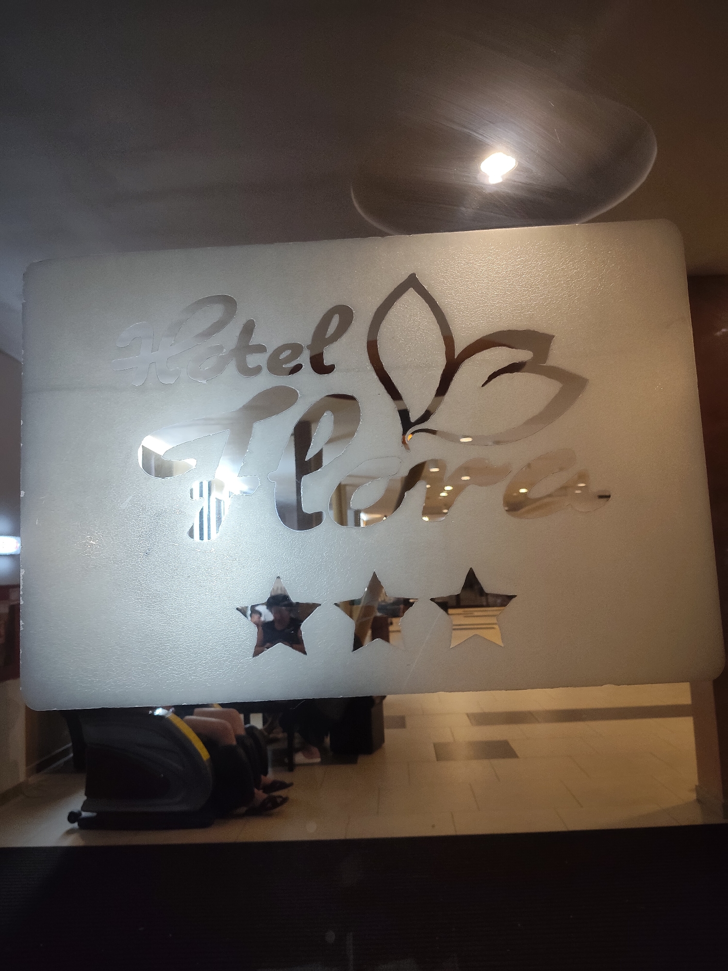 The image shows a frosted glass sign that reads "Hotel Flora" with a stylized leaf design. Below the text, there are three stars, indicating a three-star hotel. The sign is likely at the entrance of a hotel. In the reflection on the glass, a person is seen lying on their side, possibly in a suitcase or luggage. The environment appears to be an indoor public space with tiled flooring. The lighting suggests it is daytime or a well-lit interior.