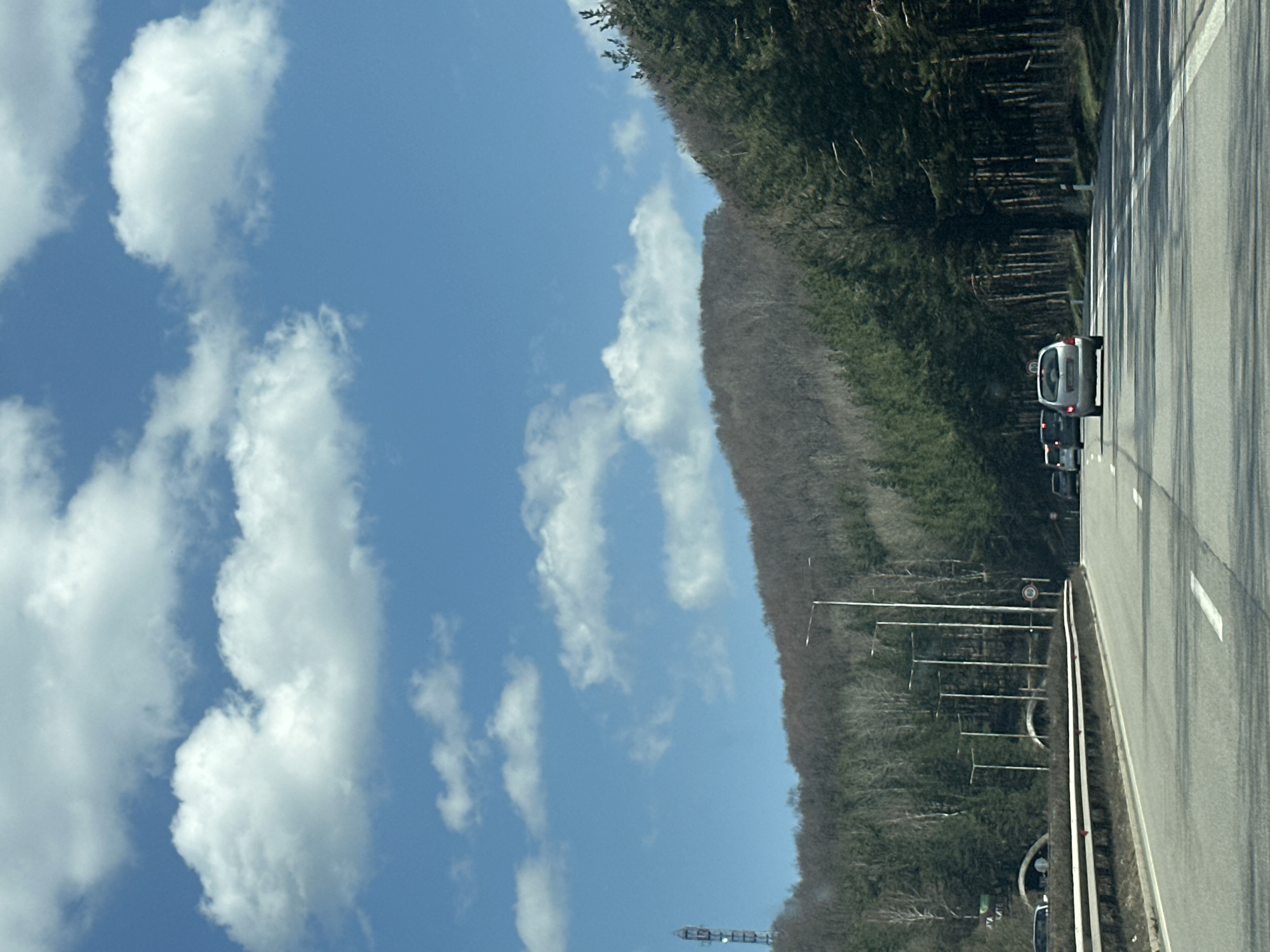 The image depicts a highway scene with a bright blue sky featuring scattered white clouds. A dense forest lines one side of the road, while a few cars are traveling on the paved highway surface. The location cannot be confirmed as Gorna Vasilitsa, Bulgaria, based solely on the image.