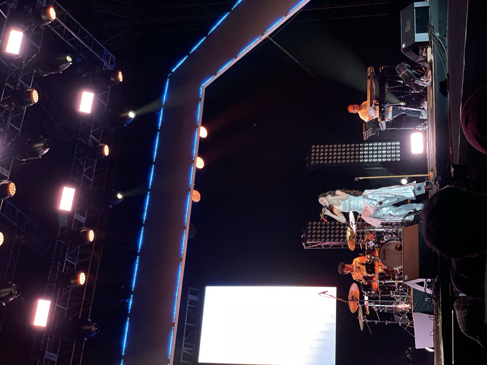 The image depicts a live music performance, possibly a concert, with a female vocalist in a sparkly silver outfit singing on stage. She is positioned above and slightly behind a drummer, who is actively playing a drum set. To the right of the singer, a keyboardist is playing their instrument. The stage setup includes a large blank screen at the bottom, a lighting rig to the left featuring multiple spotlights, and a decorative overhead structure outlined with blue LED lights. The audience is visible in the foreground, but the individuals are not fully discernible. The environment is dark, indicating an indoor venue. The city and country, Salembaran, Indonesia, cannot be determined from the image.
