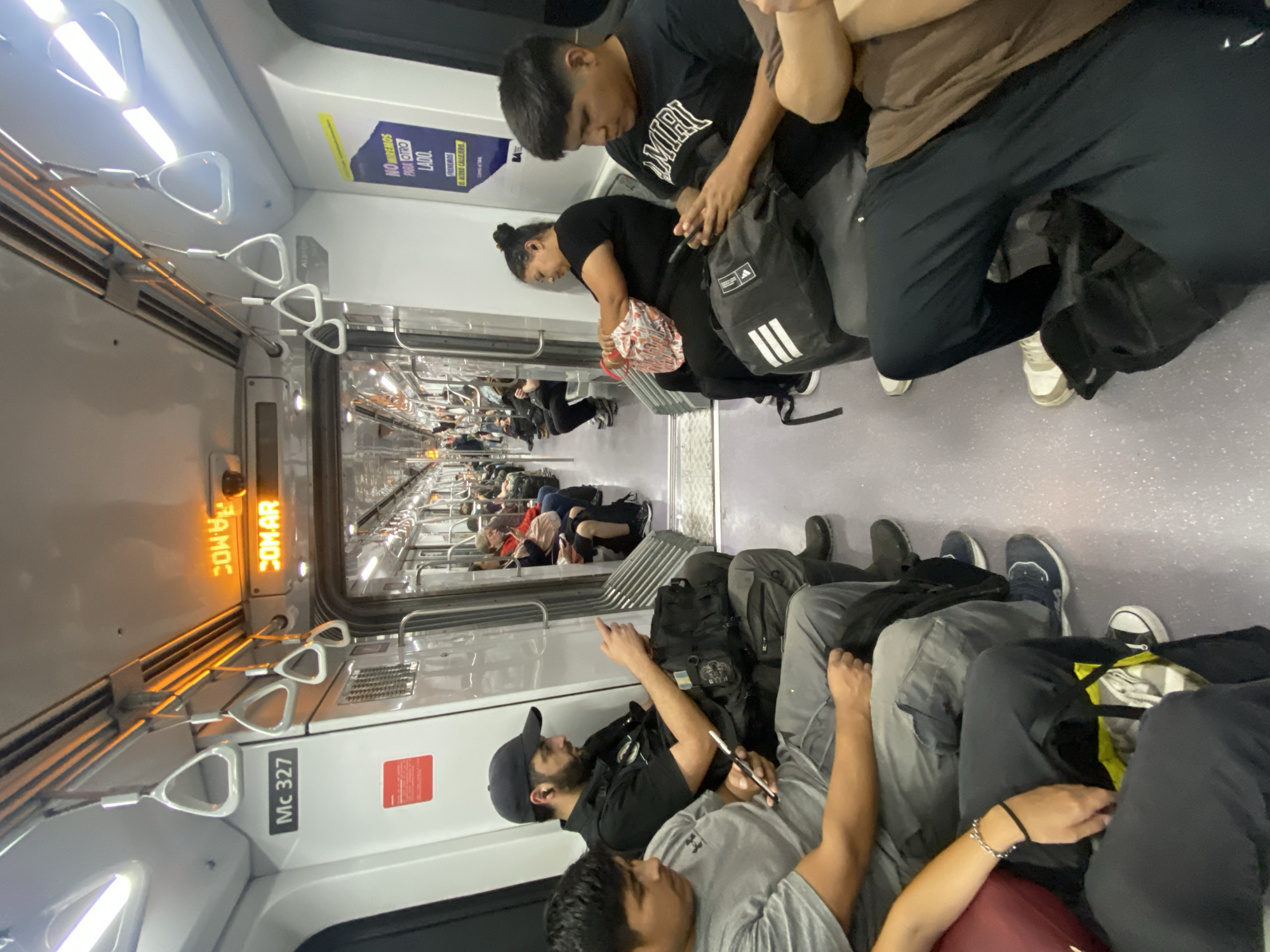 This image captures the interior of a crowded subway or commuter train car, likely in Buenos Aires, Argentina, as indicated by the Spanish text and Alstom branding, a company known for supplying trains in the region. The scene depicts numerous passengers, primarily young men and one woman, resting or sleeping on the floor and seats during transit.
The subjects are dressed casually; one man in the foreground wears a grey t-shirt, another in a black t-shirt with "AMIRI" written on it. Many have backpacks, including one with an Adidas logo, and personal belongings scattered around them. The atmosphere appears calm and weary, with individuals finding comfortable positions to rest.
The train car itself is modern and well-lit by overhead fluorescent lights. Triangular hand straps hang from the ceiling, and a long row of similar straps extends into the background. A digital display shows "COM-AR" and "PAMOC" (possibly a reflection or mirrored text). The car is identified by "Mc 327" on its side. A yellow and blue poster on the wall displays text in Spanish: "NO MIREMOS PARA OTRO LADO. FRENEMOS EL ACOSO CALLEJERO," which translates to "Let's not look the other way. Let's stop street harassment." The word "ALSTON" is also visible, likely referring to Alstom, a major train manufacturer. The scene suggests a long journey or perhaps a late-night/early-morning commute where passengers are utilizing the space to rest.