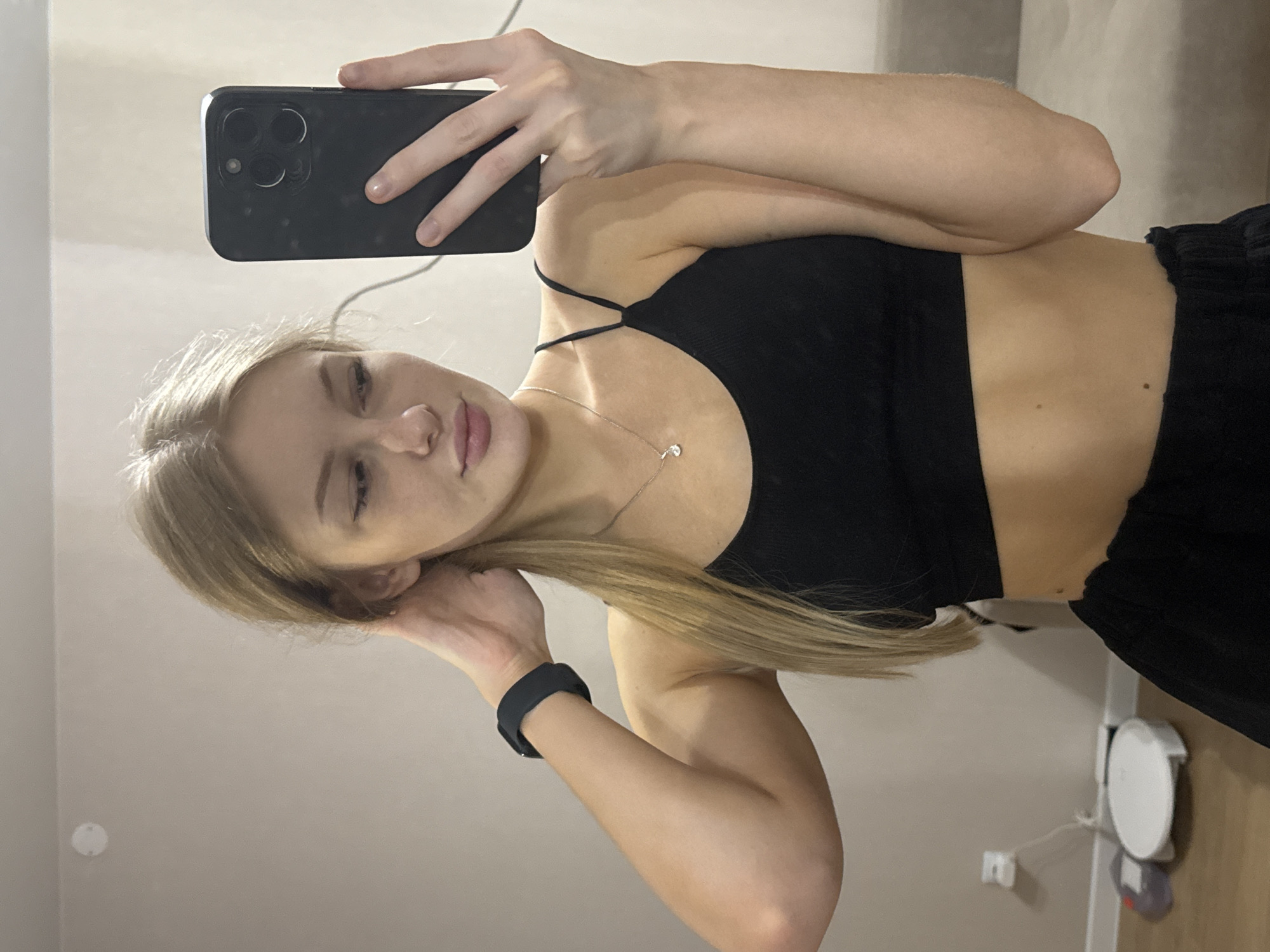 A young woman with blonde hair pulled back in a ponytail takes a selfie in a dimly lit room. She is wearing a black sports bra and black pants. Her right hand is holding a black smartphone, and her left hand is touching her hair. A black smartwatch is visible on her left wrist. The background shows a light-colored wall, a light-colored couch, and a white robot vacuum cleaner on the floor. The image does not provide any information that would allow for the determination of its location in the city of Perm, Russia.