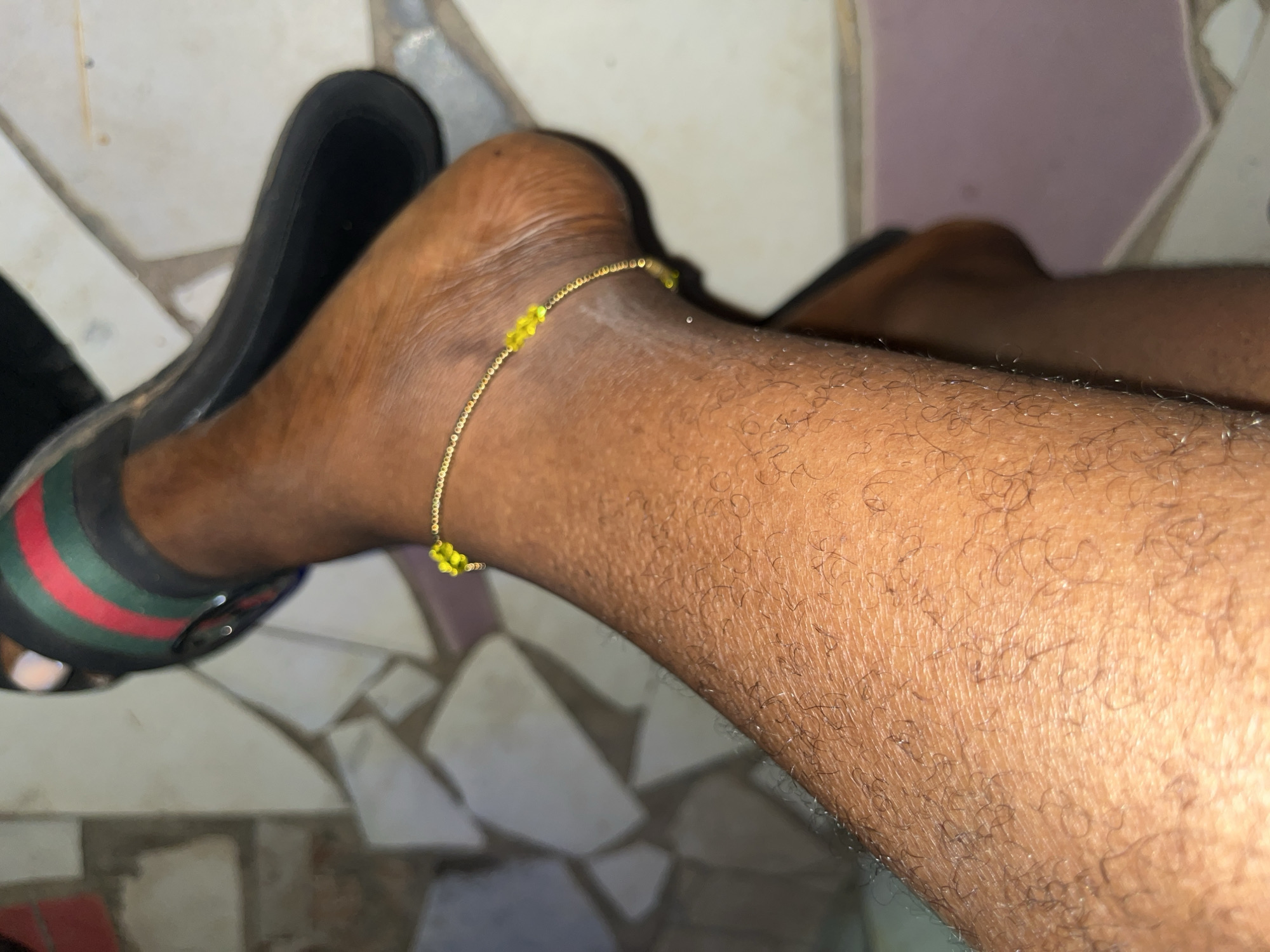 The image shows a close-up view of a person's leg and foot wearing a black sandal with red and green stripes. A delicate gold chain anklet with small yellow beads is visible around the ankle. The person's skin is dark and has visible pores and hair. The background appears to be a tiled floor with irregular geometric patterns in beige, white, and grey. The lighting suggests an indoor setting. The image does not provide any specific visual cues to identify its location within Abuja, Nigeria.