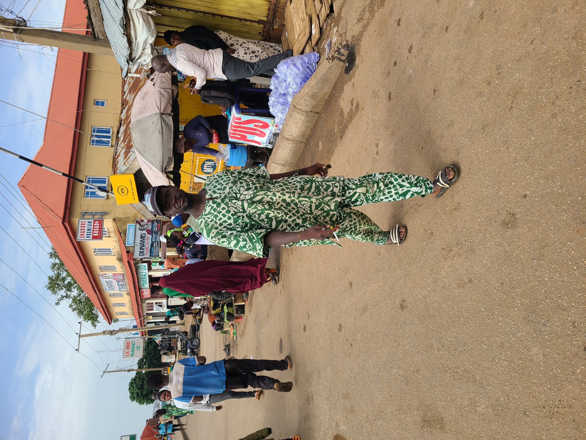 The image captures a street scene in Gura Top, Nigeria, featuring a man in a green and white patterned outfit walking on the right side of the frame. He is wearing a small white cap and sandals. He is smoking. The street appears to be paved. In the background, a multi-story building with a red roof and yellow walls is visible. Storefront signs for "SHADORC PREMIUM BREAD," "DUNAMIS," and "LA MED PHARMACY LTD." are displayed. People stand in front of a shop, and a yellow MTN-branded kiosk is present. Other individuals are walking on the street, including a group of people in blue and white shirts on the left. The sky is light blue with minimal cloud cover.