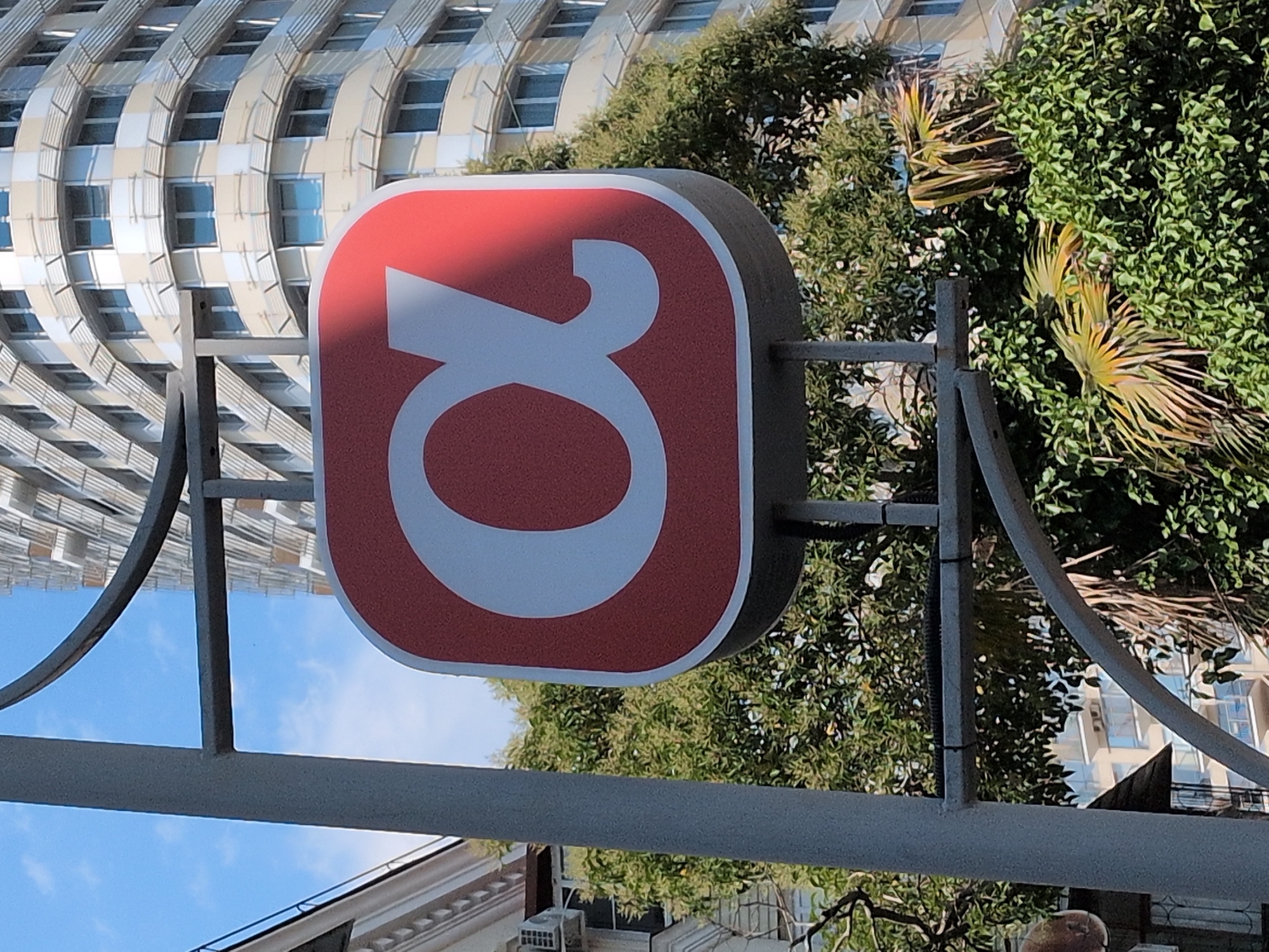 An exterior shot shows a red and white sign with the Greek letter alpha on it, mounted on a metal pole in front of a building in Sochi, Russia. The building appears to be a modern high-rise with numerous windows. Trees and foliage are visible in the foreground and around the building. The sky is blue with some clouds.