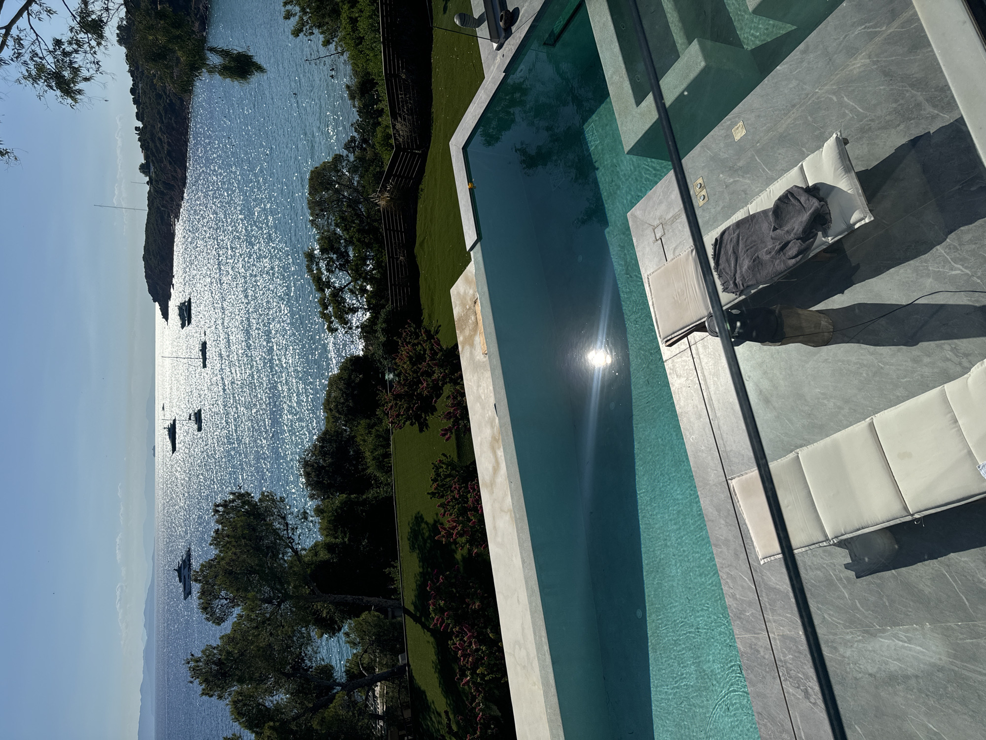 The image depicts a luxurious outdoor setting with a swimming pool overlooking a scenic bay in Vouliagmeni, Greece. The pool, a prominent feature in the foreground, is rectangular and filled with clear turquoise water, reflecting the bright sunlight. Two white sun loungers are positioned on the pool deck, one with a dark gray throw draped over it. The deck itself is paved with large, light gray stone tiles.
Beyond the pool, a vibrant green lawn slopes down towards a line of trees and lush vegetation. In the distance, the shimmering Aegean Sea stretches out, dotted with several white yachts and smaller boats. The coastline is visible, characterized by rocky outcrops and dense greenery. The sky is a clear, bright blue, with a few wispy clouds on the horizon. The sun, high in the sky, casts a strong glare on the water, creating a dazzling effect. The overall impression is one of a serene and upscale vacation destination.