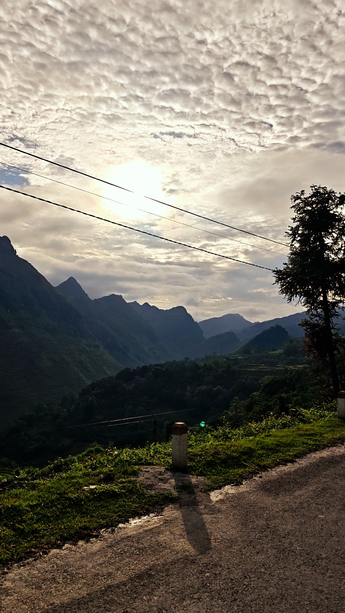 The image depicts a landscape in Lũng Cú Commune, Vietnam, featuring a mountain range under a cloudy sky. A paved road occupies the foreground, with a concrete post casting a shadow on its surface. To the side of the road, there is green vegetation. A tall tree stands near the edge of the road. Power lines are visible across the top portion of the frame, and the sun is obscured by the clouds, creating a diffused light effect. The mountains recede into the distance, their peaks silhouetted against the sky. The middle ground is filled with lush greenery.