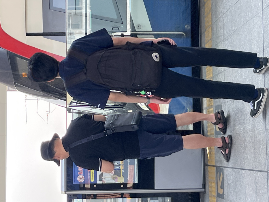 The scene depicts two individuals on an elevated train platform in Samut Prakan, Thailand. A modern train, characterized by red, white, and blue striped livery, is partially visible on the right, adjacent to the platform edge. The platform floor is paved with light grey tiles.
On the left, an adult male is seen from behind. He wears a dark bucket hat, a black short-sleeved polo shirt with white trim on the sleeves, dark shorts, and dark sandals. A dark messenger bag with a shoulder strap is slung across his body. He is positioned in front of a vertical digital screen or ticket machine that displays Thai text and appears to be interacting with it.
The second individual stands centrally, viewed from behind. This person wears a dark blue short-sleeved collared shirt, black trousers, and black sneakers with white soles, resembling Vans Old Skool footwear. A black backpack with a white Converse All-Star logo on the lower right pocket is worn. A keychain, featuring multiple green items and a peace sign charm, is attached to a strap of the backpack. A dark-colored smartphone is held in the individual's right hand. Both individuals face in the direction of the train or digital screen.