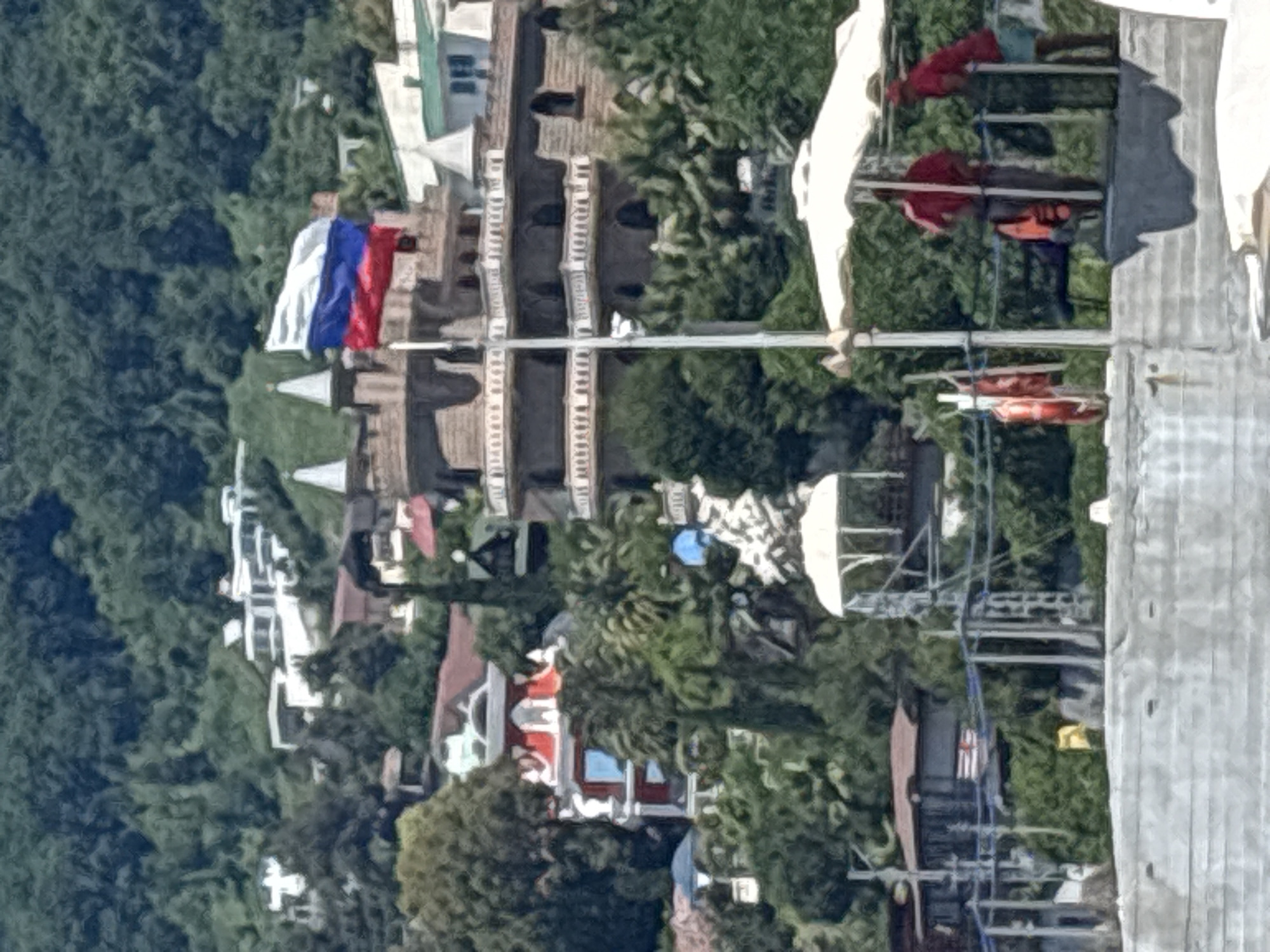 The image captures a view of Sochi, Russia, from a high vantage point, likely a cable car or ski lift. The scene is dominated by lush green trees covering a hillside, interspersed with buildings that appear to be a mix of residential homes and possibly tourist facilities. Several of the buildings have red-tiled roofs, and some are painted white or have white architectural accents.
A prominent feature is a Russian flag, with its white, blue, and red horizontal stripes, flying from a flagpole. In the foreground, part of a cable car system is visible, including its structure and what appear to be cabins or gondolas. Some life preservers, orange and red in color, are also visible, attached to the cable car infrastructure, suggesting this is a tourist attraction or area where water activities might occur.
The time of day appears to be daytime, with clear, bright lighting casting shadows, indicating sunny weather. The overall impression is one of a scenic, somewhat developed, natural landscape, typical of a resort town. There are no discernible people in the image, and no visible text. The image's orientation is vertical.