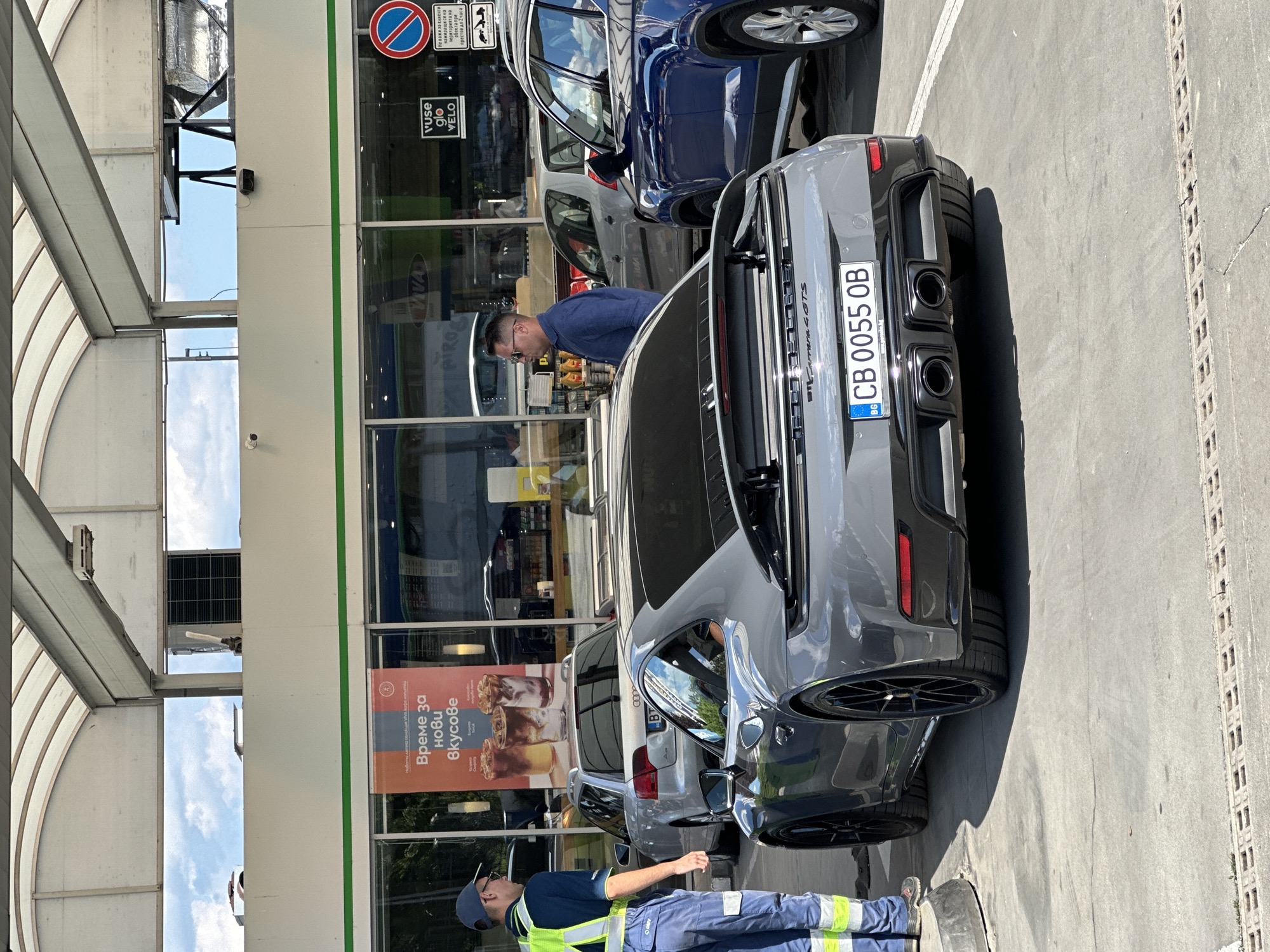 The image shows a gray Porsche 911 Carrera 4 GTS parked at a fuel station in Veliko Tarnovo, Bulgaria. A gas station attendant wearing a high-visibility vest and blue work pants stands to the left of the Porsche, facing it. To the right of the Porsche, a man in a blue shirt and sunglasses stands, looking towards the car. Behind the Porsche, an Audi sedan and a blue SUV are visible, also parked at the station. The fuel station has a covered canopy with exposed metal beams and a white ceiling. A large banner advertising coffee is displayed on the storefront window. Signs indicating "vuse glo VELO" and a no-parking symbol are also visible on the building. The ground is concrete, with some drainage grates. The sky is cloudy. The license plate of the Porsche reads "CB 0055 OB".