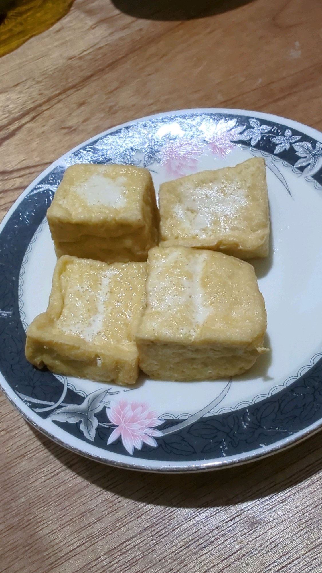 Four pieces of fried tofu are arranged on a decorative plate with a floral and leaf pattern. The plate has a dark blue and white border with metallic silver accents. The tofu pieces are golden brown and appear slightly oily, with lighter, creamy-colored interiors visible on their surfaces. The plate is placed on a light brown wooden surface with a distinct grain. The background is blurred, suggesting the image was taken in a home or restaurant setting. The scene is static and shows no people or actions. The location is not determinable from the image.