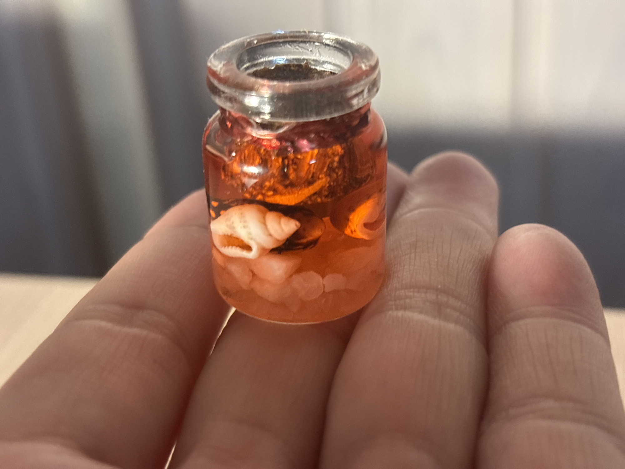 A close-up shot depicts a small, open glass jar containing what appears to be an orange-colored liquid, small white pebbles, and a seashell. The jar is being held in someone's hand, and the background is blurred, showing a glimpse of a wooden surface and a gray wall. The image provides no discernible information relating to the city of Zagatuy, Russia.