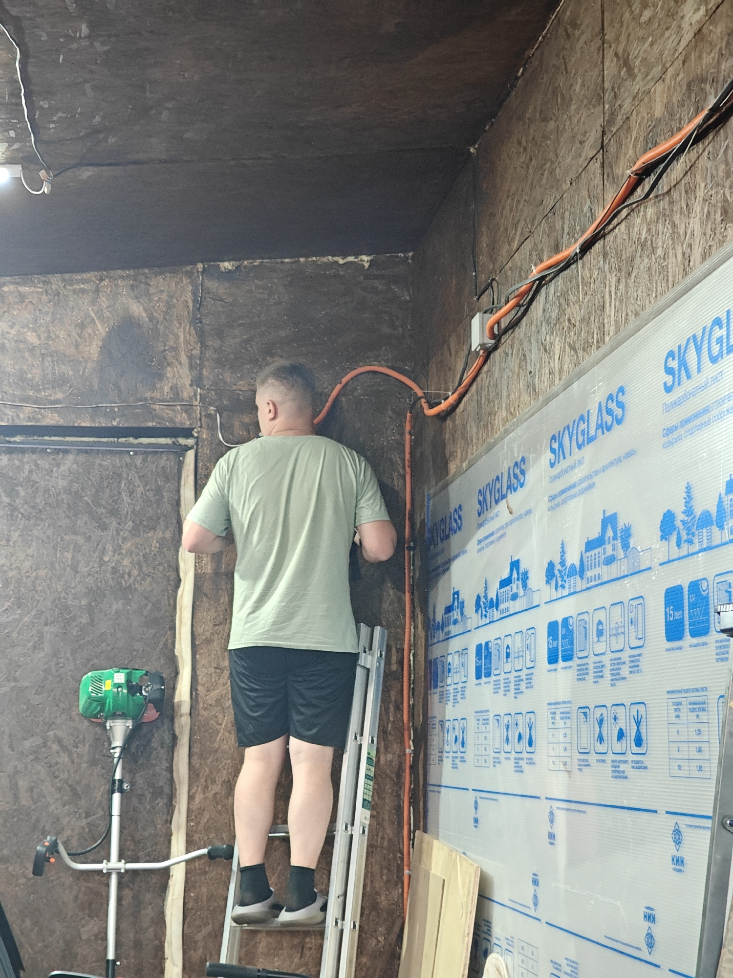 A man wearing a green t-shirt and black shorts stands on a ladder, facing away from the camera. He is in a workshop or garage, with unfinished wood paneling on the walls and ceiling. Electrical conduit, orange in color, runs along the wall and ceiling. A large sheet of clear, plastic material with blue graphics, possibly for construction, is leaning against the wall to the right of the man. A green weed trimmer is visible on the left side of the image. The location is identified as Русский Вожой, Russia.
