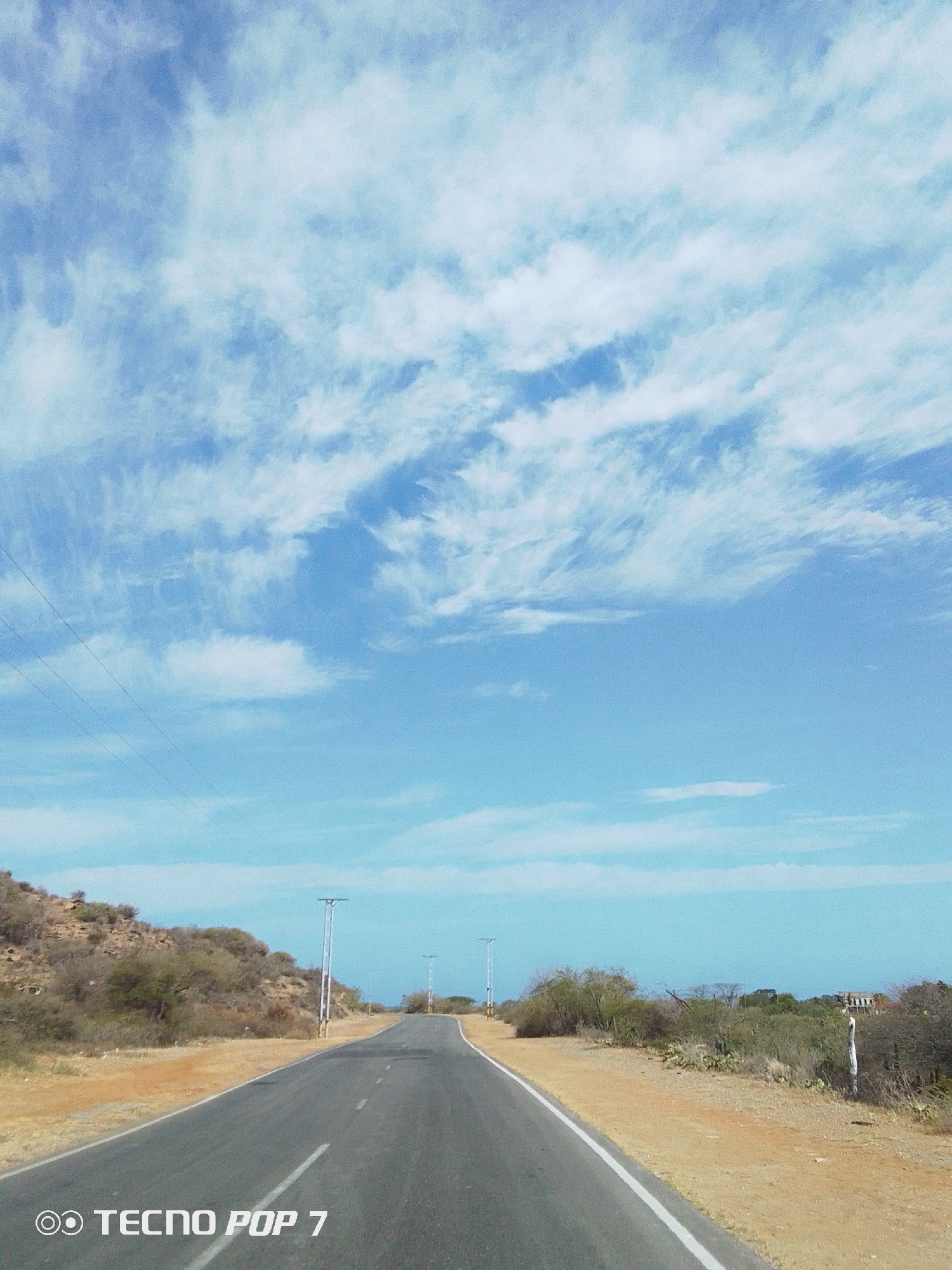The image shows a road in Pedro González, Venezuela, flanked by dry vegetation. The road, paved and marked with a white line, stretches into the distance. The sky above is blue with scattered white clouds. Power lines run parallel to the road on the left side. Utility poles are spaced along the road. The vegetation is sparse and appears arid, with low shrubs and cacti. The overall scene suggests a rural or undeveloped area. The quality of the image indicates it was taken with a "TECNNO POP 7" phone.
