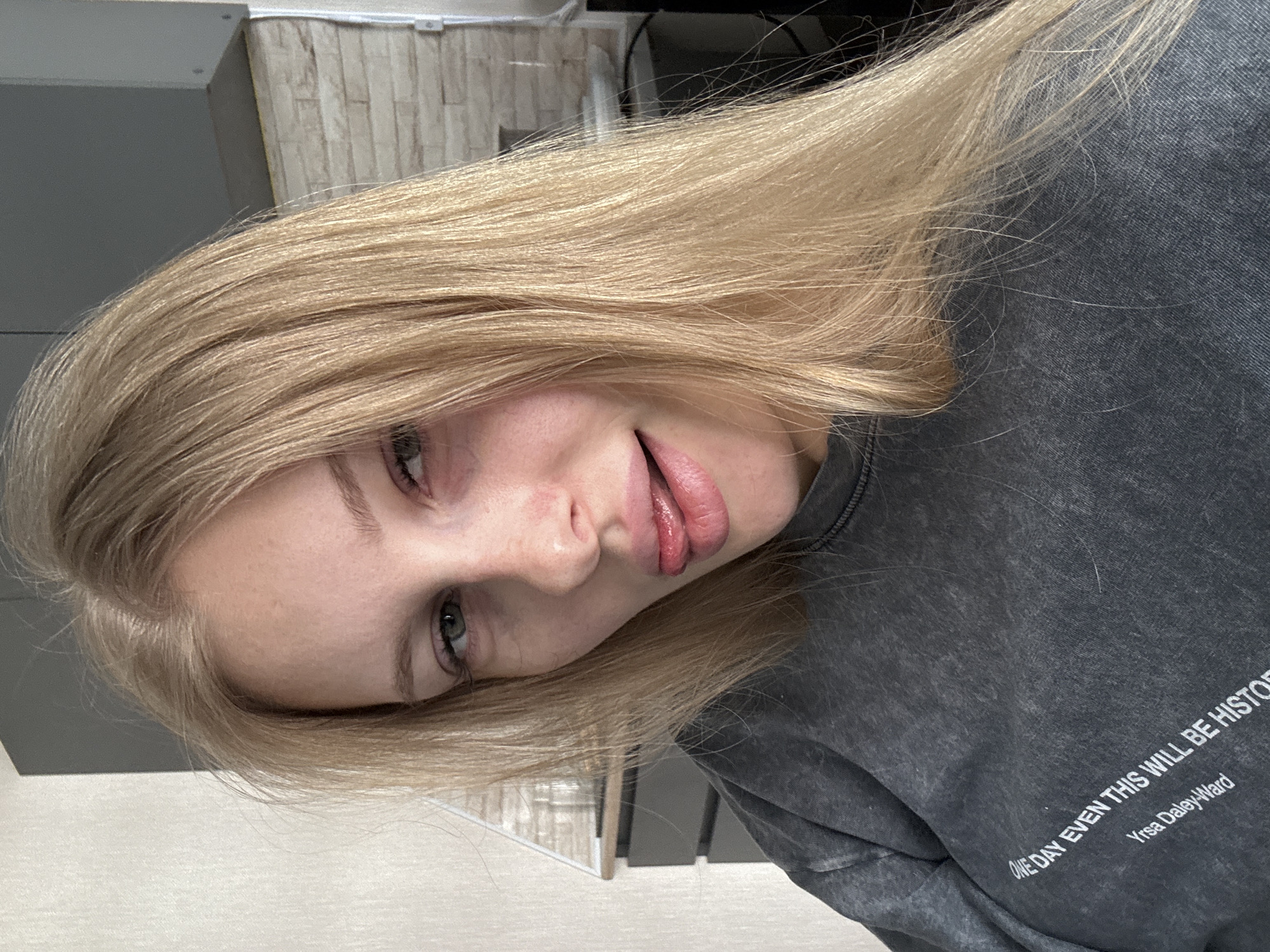 A young woman with long blonde hair, wearing a grey t-shirt with text printed on it, poses for a selfie. Her tongue is sticking out slightly. The background appears to be an indoor setting, possibly a kitchen or living area, with grey cabinetry and textured walls visible. The image does not provide any specific clues or context to indicate the location is the city of Perm, Russia.