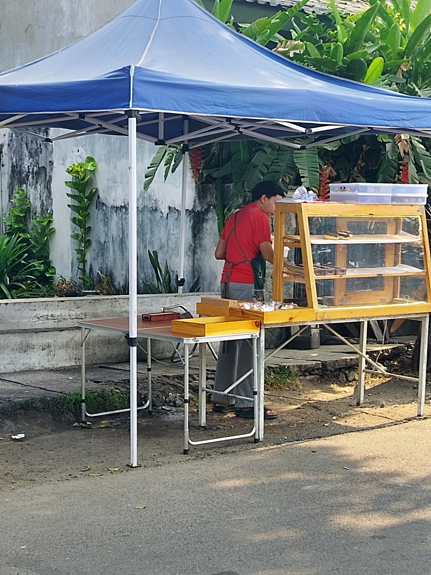 A person in a red shirt and green apron is working at a food stall. The stall is set up under a blue canopy tent. A glass display case sits on a table, holding various food items, including what appear to be pastries or cakes on shelves. Another table to the left has a wooden box and some smaller food items. The stall is situated outdoors, adjacent to a road and a wall covered in plants. The background shows lush green foliage. This scene is located in Mojokerto, Indonesia.