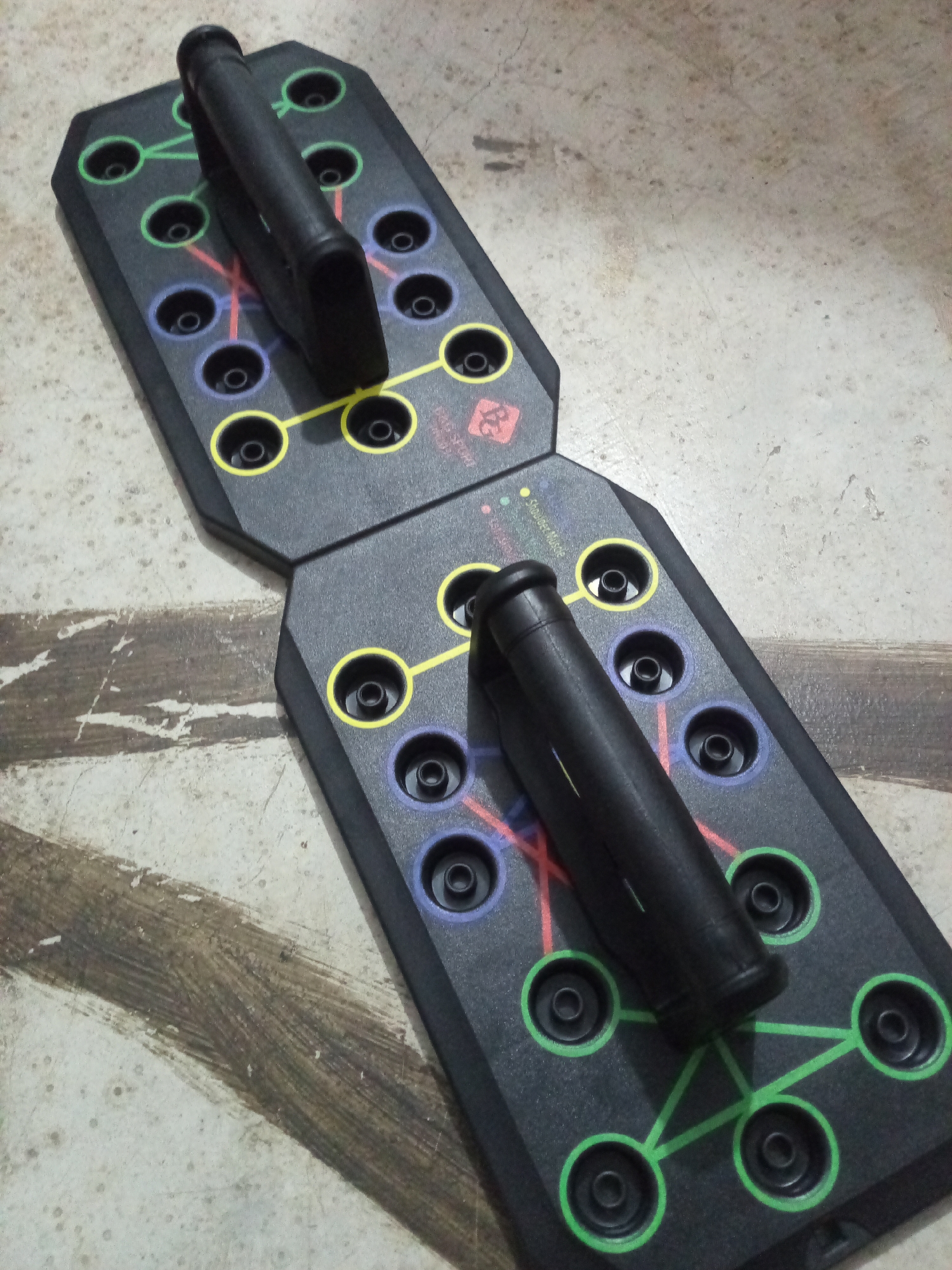 A push-up board is lying on a gray surface. The push-up board is black and divided into two halves, connected in the middle. Each half has multiple holes, each surrounded by a circle of a different color: green, red, yellow, and blue. Black handles are inserted into the holes. The image appears to have been taken indoors in Davao City, Philippines.