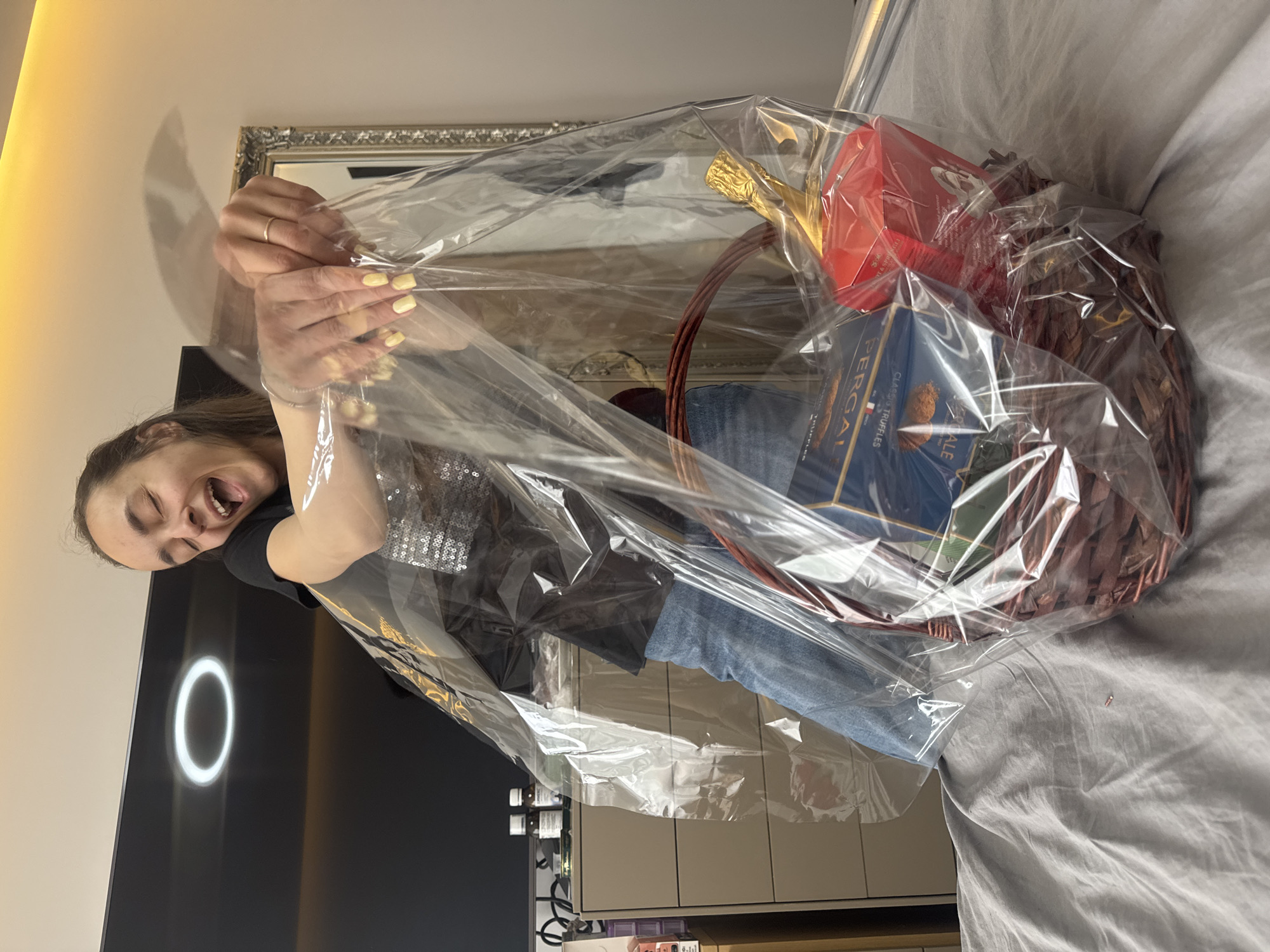 A woman with brown hair, wearing a black top with glitter accents and blue jeans, is holding a cellophane-wrapped gift basket. The gift basket contains a gold-topped bottle and boxes. The basket rests on a gray sheet, possibly a bed. The background includes a wall, a mirror, a black TV with a ring light mounted on it, and a cabinet. The image appears to have been taken indoors. There is no information about the location of the city of Sofia, Bulgaria.