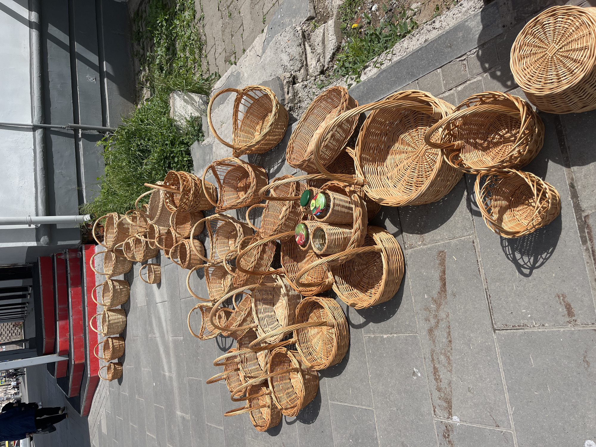 The image shows a collection of wicker baskets of various sizes and shapes displayed on a gray paved surface. The baskets range from small to large, some with handles and others without. There are some baskets that contain colorful jars of pickled goods. The location appears to be outdoors, with a stairway and building visible in the background. The image is taken in Izhevsk, Russia.
