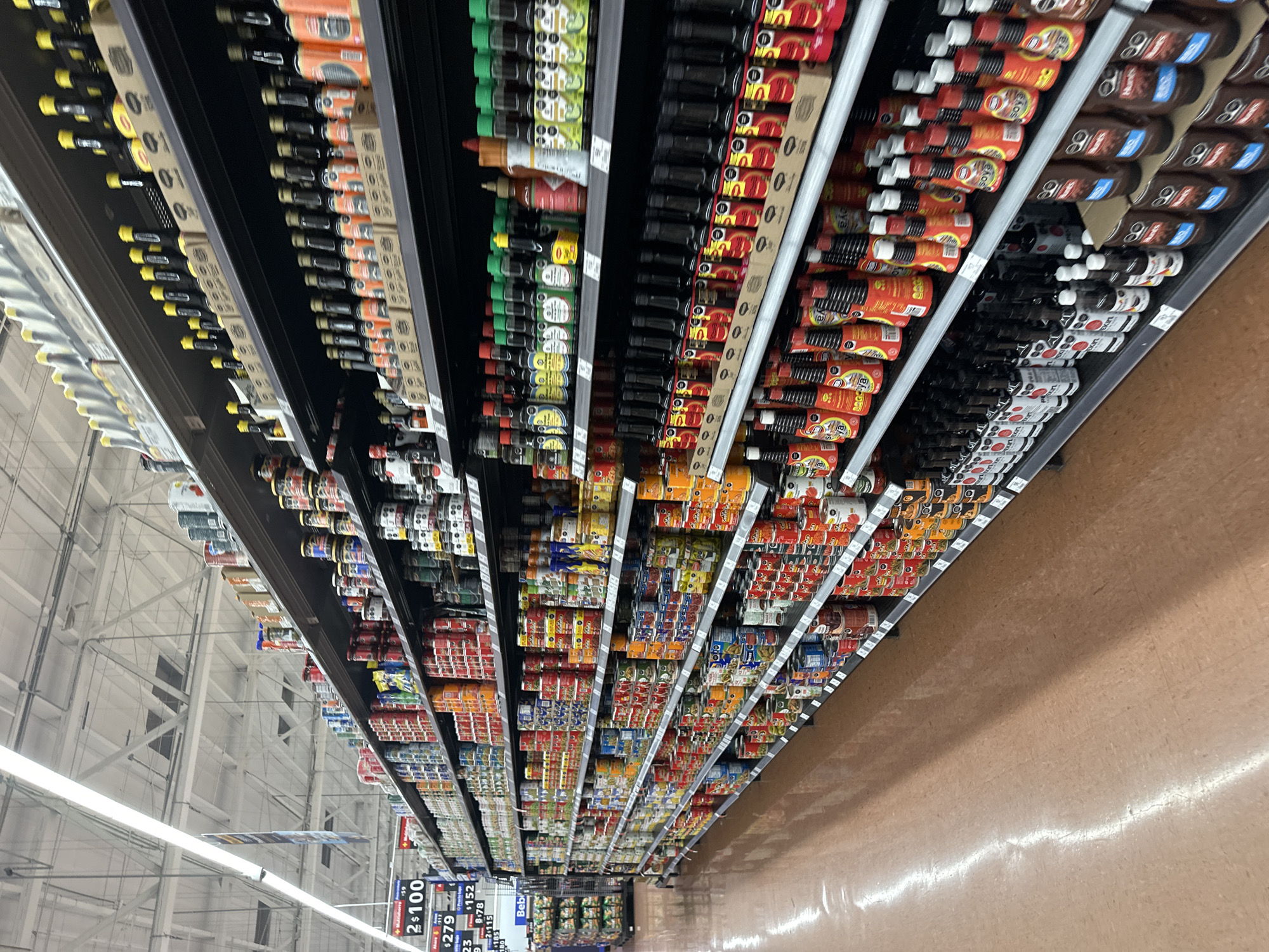 The image shows a wide-angle perspective of a grocery aisle in a store. Rows of shelves are stocked with various food products, including sauces, condiments, and canned goods. The shelves extend into the distance, creating a sense of depth. The floor is a light brown color and appears polished. Price tags are visible on the shelves, indicating the cost of the items. The location is Huixquilucan de Degollado, Mexico.