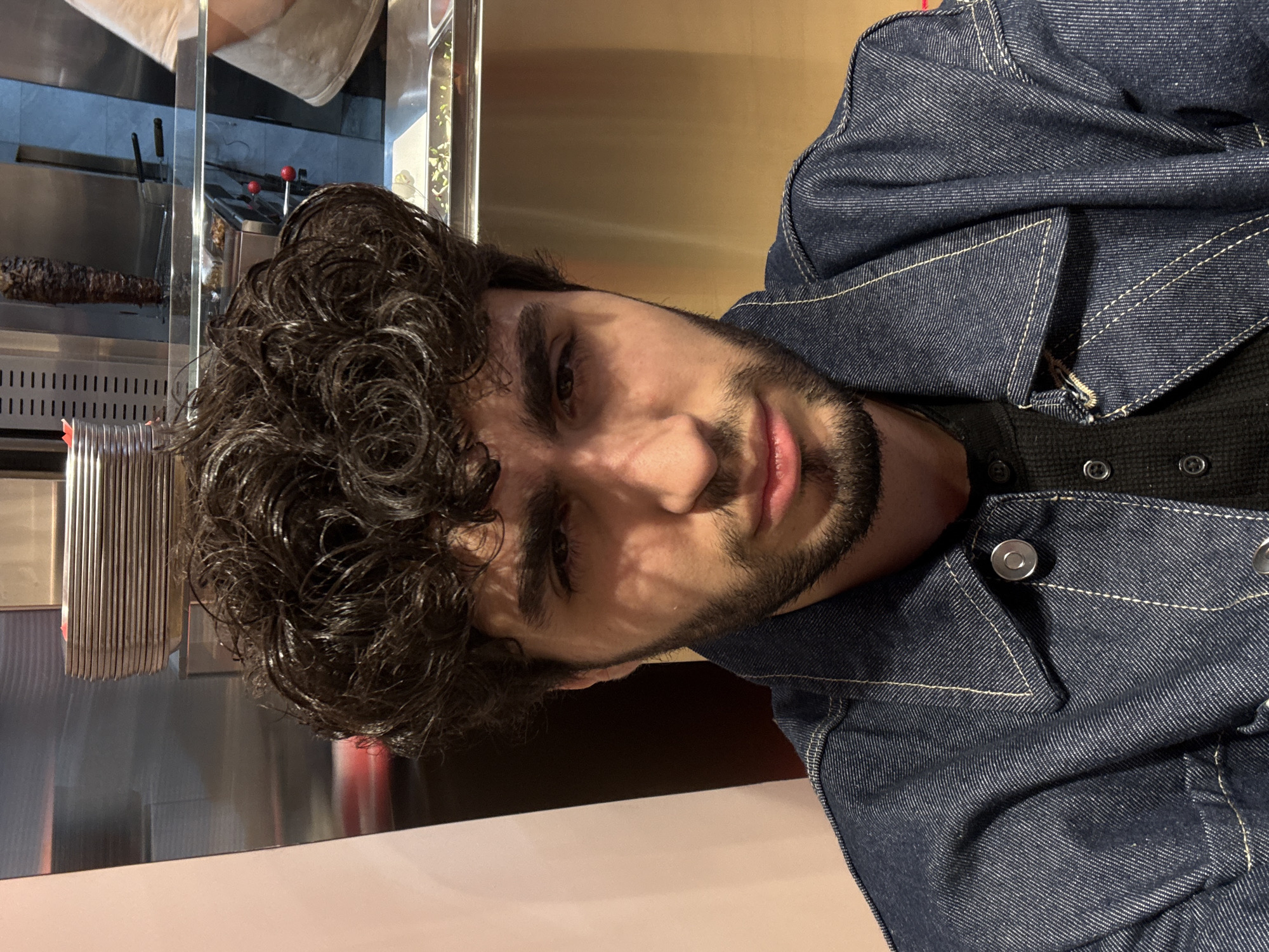 The image, when correctly oriented, presents a close-up portrait of a man positioned on the left side of the frame, looking directly towards the viewer. He has dark, curly hair and a short, dark beard. He is wearing a dark blue denim jacket with prominent light-colored stitching, worn over a dark, possibly waffle-knit, shirt. His expression is neutral to slightly contemplative, with his lips slightly parted. Shadows from an overhead or angled light source fall across his face, emphasizing his features.
The background, partially blurred and seen through a reflective surface behind the man, reveals an indoor commercial kitchen or food preparation area. A prominent detail in the background is a large skewer of roasted meat, likely kokoreç or a similar dish, slowly rotating on a vertical spit over a heat source. Surrounding this are stainless steel surfaces, various kitchen equipment, and some tools with red handles. The setting suggests an eatery, restaurant, or food stall in Yerevan, Armenia. There is no visible text within the image.