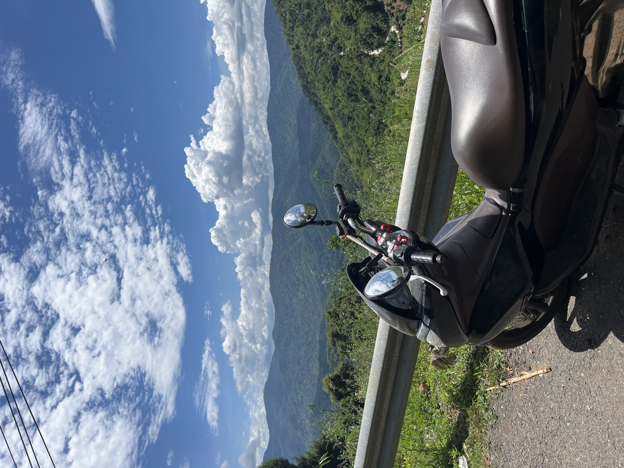 A black scooter is parked on the side of a road overlooking a lush green mountain landscape under a blue sky with scattered white clouds. The scooter is positioned in the foreground, taking up the right half of the image, with its brown seat and handlebars visible. A metal guardrail runs along the edge of the road. The mountainous terrain stretches into the distance, with a winding road visible on one of the slopes. The scene is bathed in sunlight, casting shadows on the road. The image appears to have been taken on a sunny day in Son Dien, Vietnam.