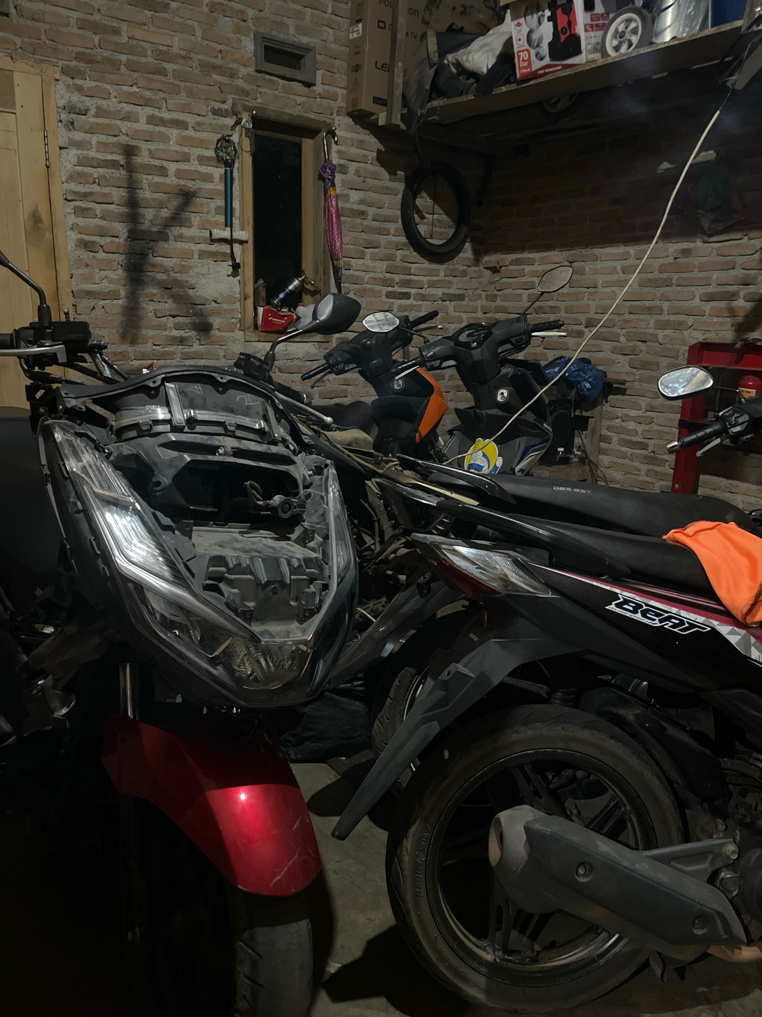 The image depicts a dimly lit garage or workshop, likely in Kebumen, Indonesia, filled with several motorcycles. In the foreground, a red and black motorcycle is partially disassembled, with its front fairing removed, revealing the internal components. Behind it, a black motorcycle with the "Beat" logo is visible, partly covered by an orange cloth. Other motorcycles, some with orange accents, are also present in the background. The walls are constructed of brick, and a shelf above holds various items, including a pressure washer and a spare tire. A small window and a door are visible on the left side of the brick wall. The overall impression is one of a working space for motorcycle maintenance or repair.