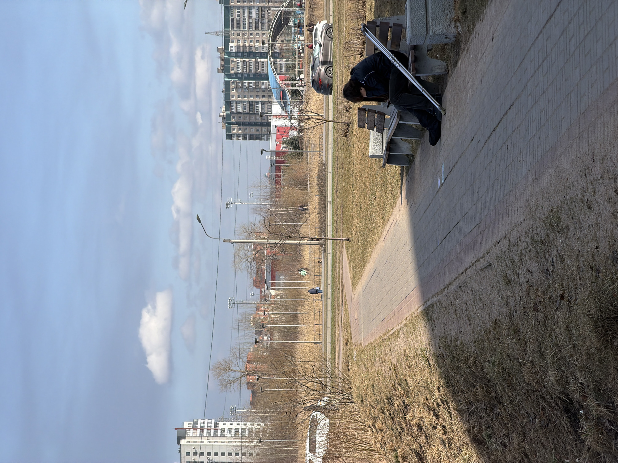 The image depicts an outdoor urban scene in Saint Petersburg, Russia, specifically near the Sports Complex "Kolomyagi" in the Kolomyagi municipal okrug. The scene is illuminated by natural sunlight under a partially cloudy blue sky, characteristic of late winter or early spring, with predominantly dry, light brown grass and bare deciduous trees.
In the foreground, a paved pathway made of reddish-brown and gray bricks extends diagonally from the lower right to the mid-left. To the right of the path, a person is seated on a park bench with concrete supports and wooden slats. The individual is wearing a dark jacket and dark pants, with their head bowed and obscured by hair, facing towards the right side of the frame. A silver crutch leans against the bench next to the seated person. The ground surrounding the pathway and bench consists of dry, sparse grass and visible soil.
The midground features a wide expanse of dry, yellowish-brown grass and scattered leafless bushes and small trees. Beyond this open area, a paved road is visible with several vehicles, including a dark brown SUV and a lighter-colored car. A metal fence runs parallel to the road. Further back, a series of tall streetlights with multiple fixtures delineate an open field. Several indistinct human figures are visible walking in the distance on what appears to be a parallel path or sidewalk. Prominently in the midground-background are the red building and a structure with a distinctive blue arched roof, identifying features of the Sports Complex "Kolomyagi".
The background is dominated by various multi-story residential buildings, primarily in shades of grey and light green, consistent with typical urban architecture in the region. A tall, white residential building with a red flag or antenna on its roof is visible on the far left. The sky above is bright blue with scattered white cumulus clouds.
