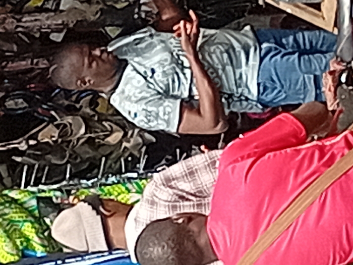 The image depicts an outdoor scene in the city of Ayangba, Nigeria, at what appears to be a vendor stall. In the foreground, positioned with their back to the viewer, is an individual wearing a red short-sleeved shirt with a brown strap diagonally across their back. Directly behind them, slightly to the left, and also facing away from the viewer, is another individual wearing a light-colored knit cap and a light-colored, possibly plaid, long-sleeved shirt. To the right, a third individual with dark skin and short hair stands, facing downwards and slightly to the left. This person is wearing a light grey patterned short-sleeved t-shirt and blue denim shorts or jeans, with their hands held up in front of them, palms inward, as if examining or manipulating small items. The background features a dense vertical display of dark-colored merchandise, possibly footwear or small bags, hanging from a structure. To the far left in the background, a green and yellow patterned fabric is visible. Partially obscured dark objects are present on a surface in the lower right foreground, adjacent to the individual in the grey shirt. No direct interaction between the visible individuals is clearly depicted.