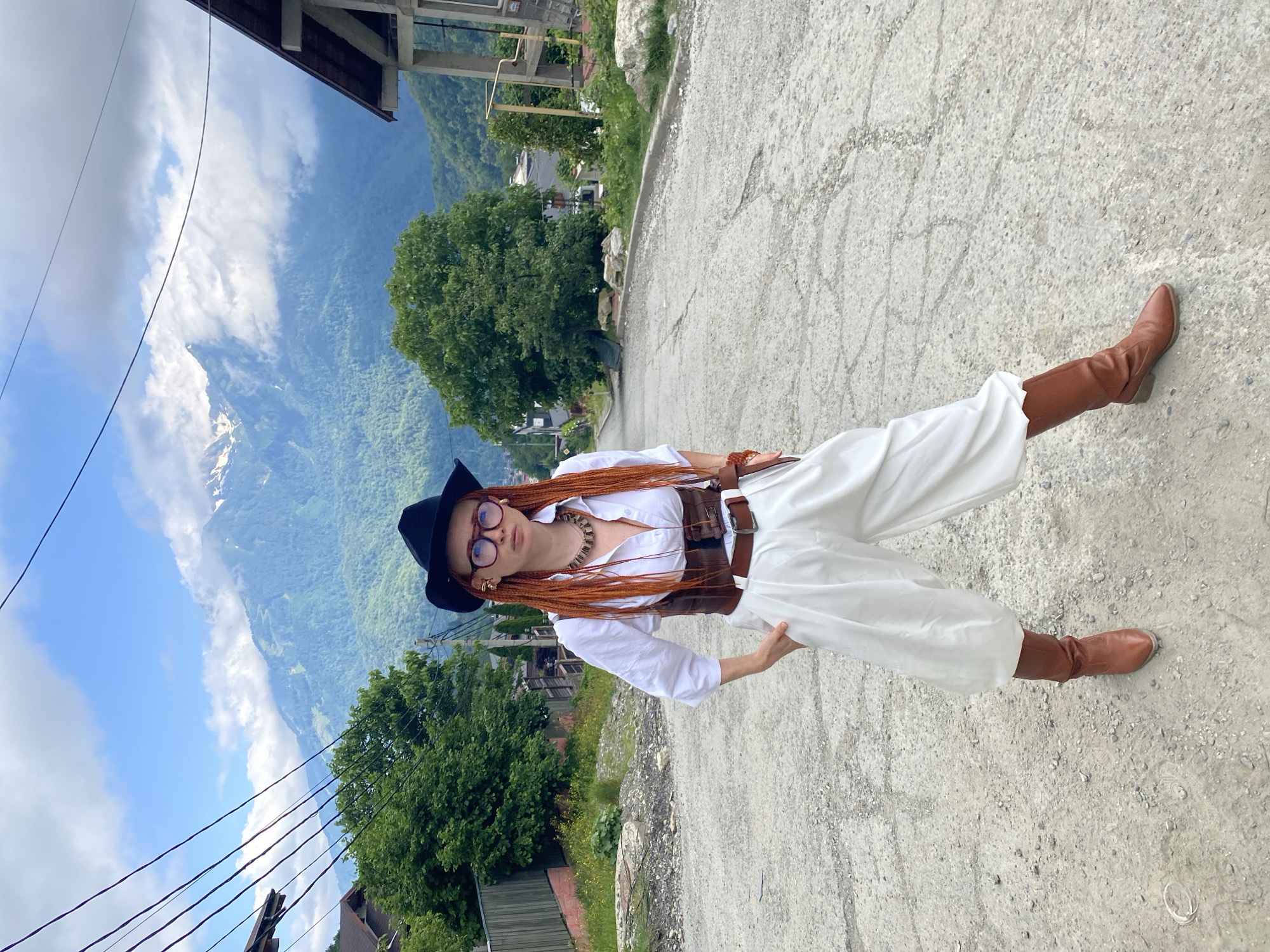 In Krasnaya Polyana, Russia, a woman with bright orange braided hair stands on a sloped asphalt road. She wears a black hat, large round glasses, a white collared shirt, a brown leather corset, wide white pants, and brown boots. She has a beaded necklace and is wearing jewelry on her ears and wrists. The background features lush green mountains partially shrouded in clouds, with houses and trees dotting the landscape. Overhead power lines are visible in the foreground.