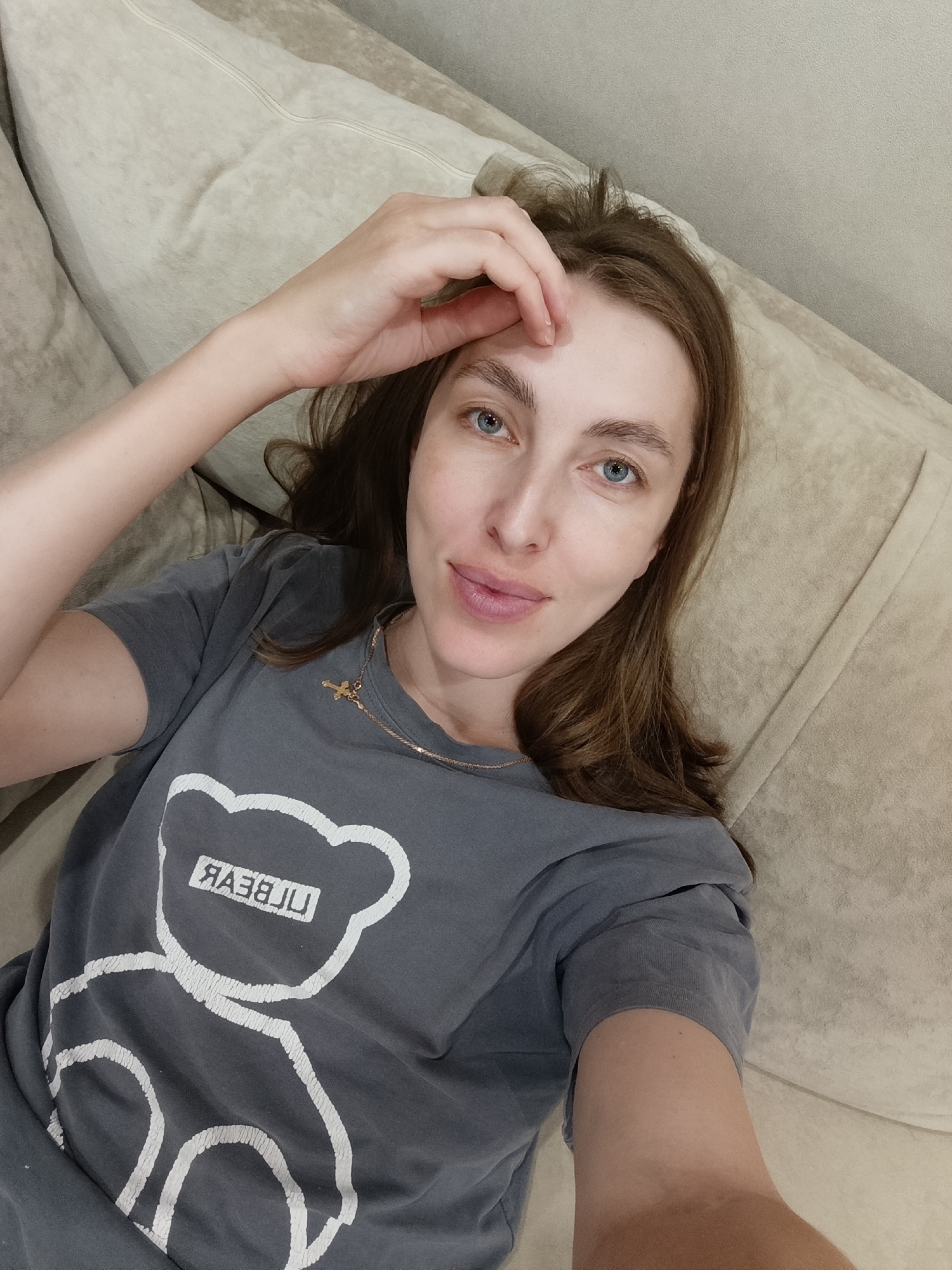 A young woman with long brown hair and blue eyes lies on a beige sofa, looking up at the camera. She is wearing a gray t-shirt with a white outline of a bear and text. Her right hand is resting on her forehead. The environment appears to be an indoor living space. The location is in the city of Chaikovsky, Russia.