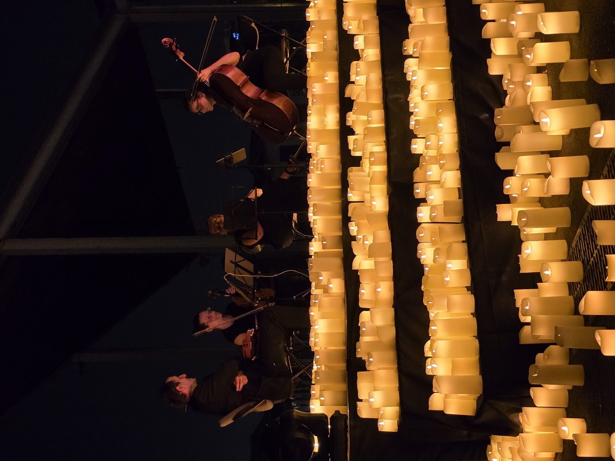 The image depicts an outdoor nighttime scene with a group of musicians playing instruments, possibly a string quartet, illuminated by rows of lit candles. The musicians, one male and three females, are seated and positioned on a raised platform or stage. The male musician on the left is wearing a suit and appears to be speaking or conducting. The female musicians are playing a violin and a cello. In the foreground, a vast array of pillar candles, all lit and emitting a warm glow, are arranged on multiple tiers, creating a luminous display that dominates the lower half of the image. The background is dark, suggesting the scene is set at night, possibly with urban structures or natural elements faintly visible. The overall atmosphere is serene and commemorative, highlighted by the soft candlelight and the focused musicians. The location is identified as Sant Adrià de Besòs, Spain.