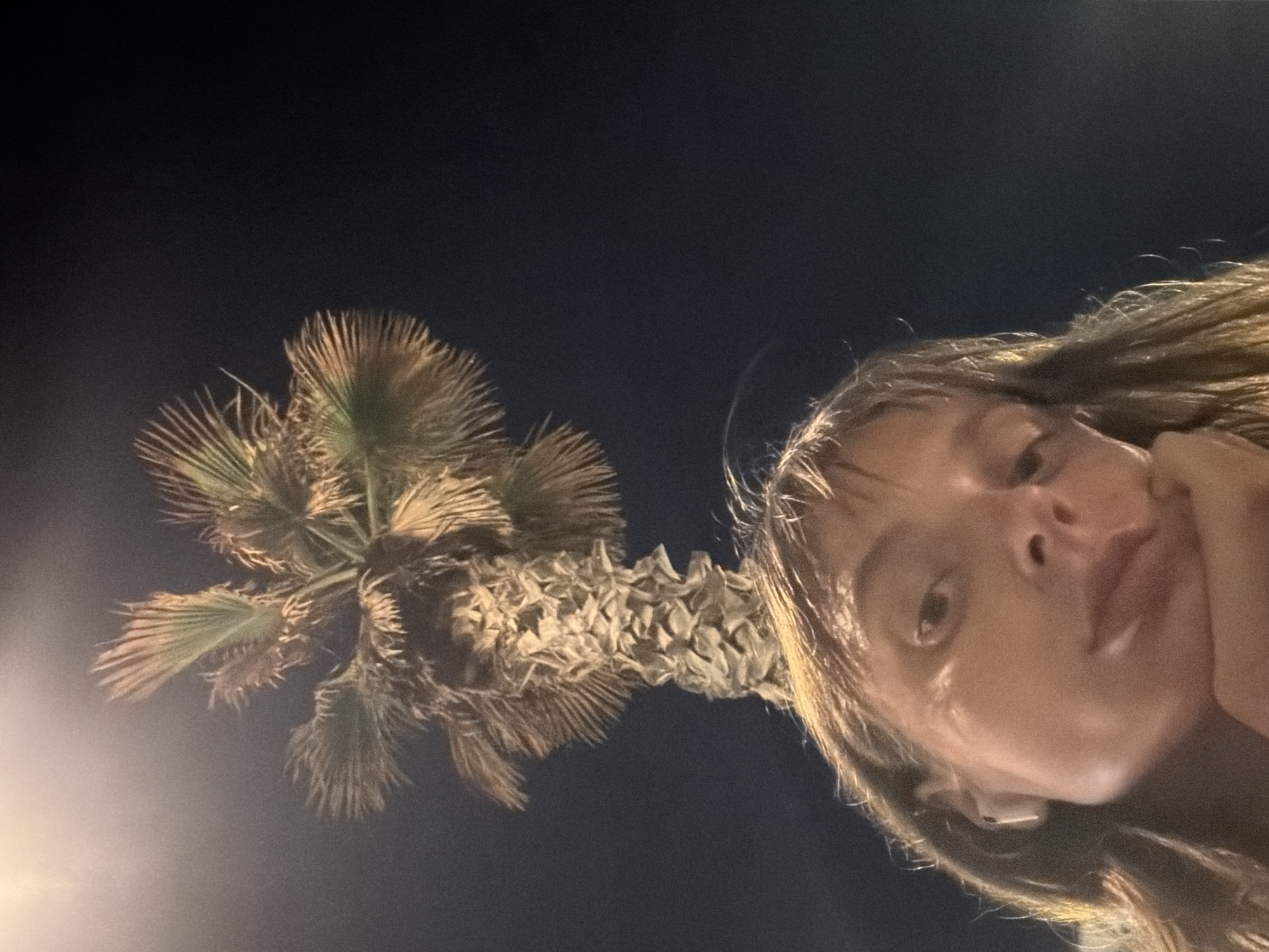 The image is a close-up, low-angle selfie of a person with shoulder-length brown hair and light-colored eyes, wearing white earbuds. The person is resting their chin on their hand and looking upwards. Behind them, a palm tree trunk and fronds are visible against a dark night sky, with a bright light source casting a glow on the upper left portion of the image. The setting appears to be outdoors at night in Tel Aviv, Israel, possibly near a street or public area given the presence of artificial light and the distinctive palm tree.