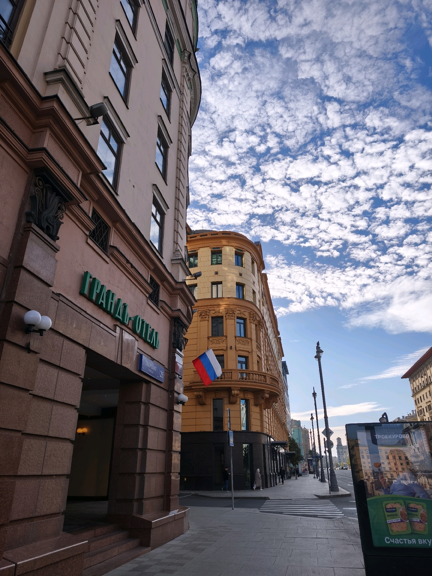 The image captures a street scene in Moscow, Russia, specifically on Tverskaya Street, indicated by the blue street sign reading "ТВЕРСКАЯ 26". The foreground is dominated by the textured, reddish-brown facade of a building with large, angular stone blocks. Above this, a sign in green Cyrillic lettering reads "ГРАНД ОТЕЛЬ" (GRAND HOTEL). Two round white lamps are mounted on the facade.
To the right of the Grand Hotel, a curved, orange building with classical architectural details, including ornate balconies and decorative stonework around windows, is visible. A Russian flag, red, white, and blue, hangs from a pole attached to this building. Further down the street, a pedestrian crossing is marked on the asphalt. A tall, black lamppost stands near the crossing.
The background shows a street lined with various buildings of different architectural styles and colors, extending into the distance. The sky above is a bright blue, dotted with numerous small, white, puffy clouds. A poster board with images and Cyrillic text is positioned on the sidewalk to the right, partially obscuring the view of the street beyond. A few pedestrians are walking on the sidewalk. The overall impression is of a city street on a clear day.
