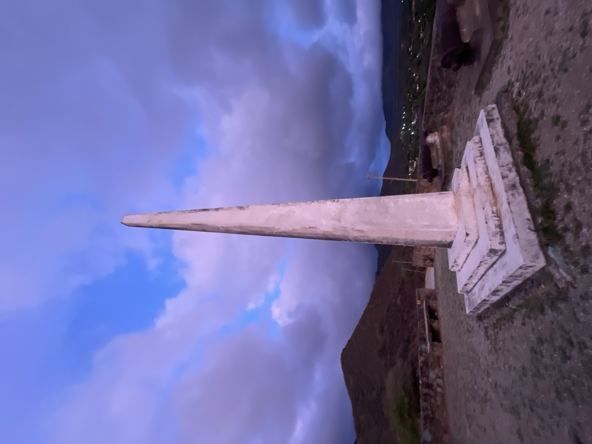 The image captures a twilight scene at a historical monument in Santa Ana, Venezuela. Dominating the foreground is a tall, slender obelisk made of weathered concrete, resting on a tiered square base. The monument is situated on a raised, rocky platform, with two dark, ancient cannons positioned on either side, suggesting a former military or defensive site.
Behind the obelisk, a low, weathered stone wall runs horizontally, punctuated by a simple concrete bench. Beyond the wall, the landscape slopes upwards into rugged, dry hills. In the distance, the lights of a town twinkle faintly against the darkening sky, hinting at the urban sprawl of Santa Ana. The sky itself is a dramatic blend of deep blues and purples, streaked with illuminated clouds, indicating either the last moments of sunset or the first hints of dawn. The overall atmosphere is serene and somewhat somber, conveying a sense of history and quiet contemplation.