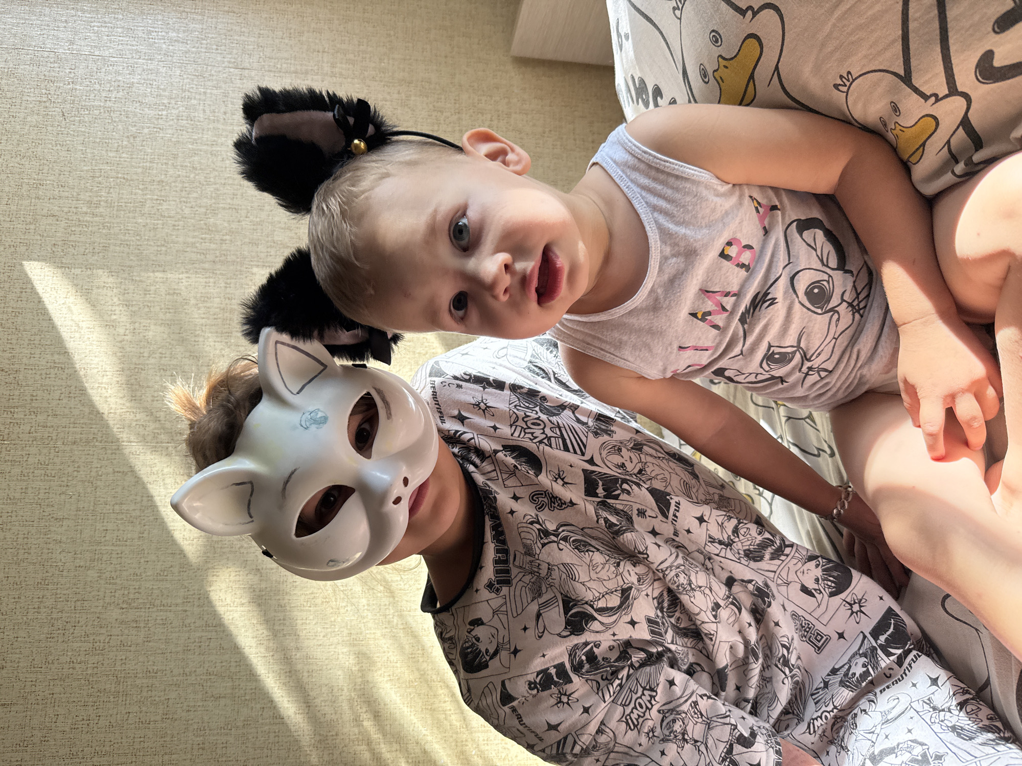 Two young individuals, one wearing a white cat mask and a graphic t-shirt, and the other a younger child with black cat ears, are seated together on a bed indoors. The older individual's face is partially obscured by the mask, while the younger child looks directly at the camera with an open mouth. The bed is covered with a duvet featuring a pattern of cartoon ducks. Sunlight streams in from the left, casting shadows on the textured wall behind them. The setting appears to be a bedroom in a residential building, possibly in Rusanovo, Russia, given the context provided.