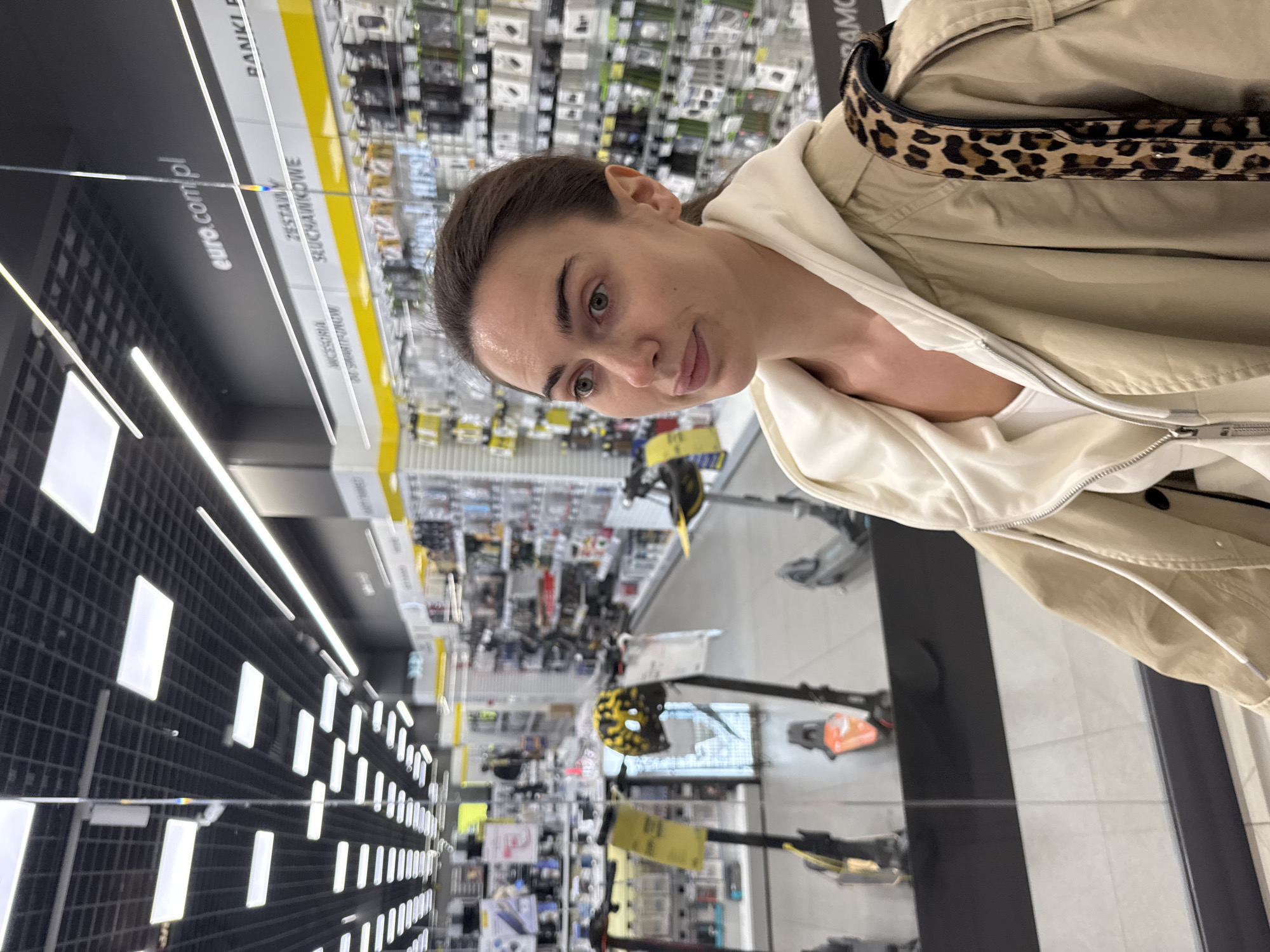The image captures a woman in an indoor retail environment, specifically an electronics store located in Warsaw, Poland. The woman, with dark hair pulled back and light-colored eyes, looks directly at the camera with a neutral to slightly amused expression. She is wearing a light beige or cream hooded jacket over a white top, with a distinctive leopard print strap or bag visible over her right shoulder.
The background is intentionally blurred, directing focus to the woman. However, elements of the store are discernible: rows of shelves filled with various packaged products, likely electronics accessories. Modern black display units with illuminated rectangular panels line the aisles. The flooring consists of light-colored tiles. Further back in the store, several electric scooters or similar personal mobility devices are on display.
Visible text on yellow signage strips confirms the store's identity and product categories. "euro.com.pl" is clearly seen, indicating the Polish electronics retailer RTV Euro AGD. Other Polish words visible include "SŁUCHAWKOWE" (headphones/earphones), "AKCESORIA" (accessories), and "DO SMARTFONÓW" (for smartphones), along with partial views of "ZESZAWY" (sets) and "RANKI" (likely part of "powerbanki" - power banks).
The scene is lit by artificial indoor lighting, making the time of day and external weather conditions undeterminable. The woman appears to be casually standing in an aisle.
