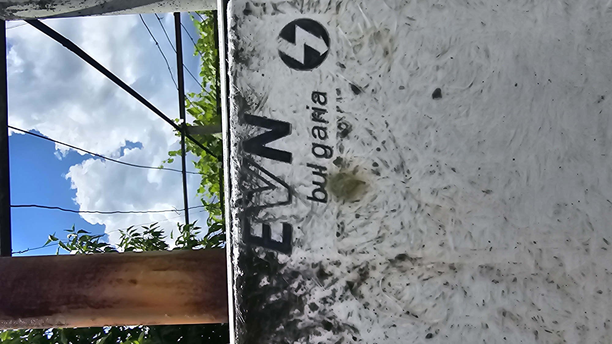 The image shows a weathered, white, textured surface with "EVN bulgaria" written in dark lettering. To the right of the text is a circular logo with a lightning bolt symbol. The surface appears to be part of an outdoor utility box or structure. In the background, there is a rusty pipe on the left, green foliage, and a bright blue sky with white clouds. The overall scene suggests an outdoor urban or semi-urban environment, possibly in Karnare, Bulgaria, given the inscription.