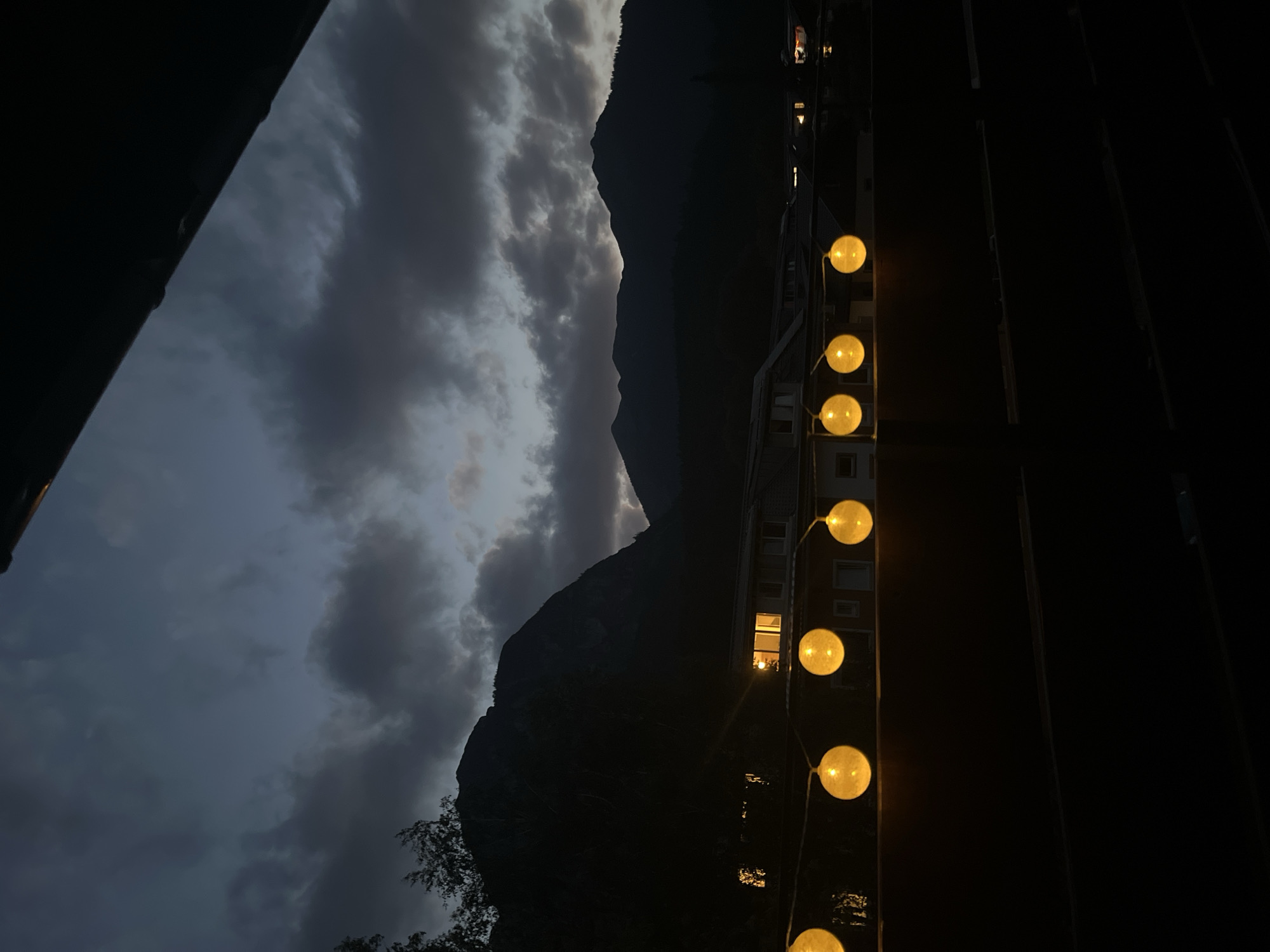 The image depicts a twilight scene in Garmisch-Partenkirchen, Germany, with a string of yellow globe lights illuminating the foreground. The lights are suspended above a dark railing, casting a warm glow against the darkening sky. Behind the lights, a row of buildings with illuminated windows is visible, suggesting a residential or commercial area. Further in the background, dark, imposing mountains rise against the cloudy twilight sky. The overall impression is one of a peaceful evening in a mountainous Bavarian town.