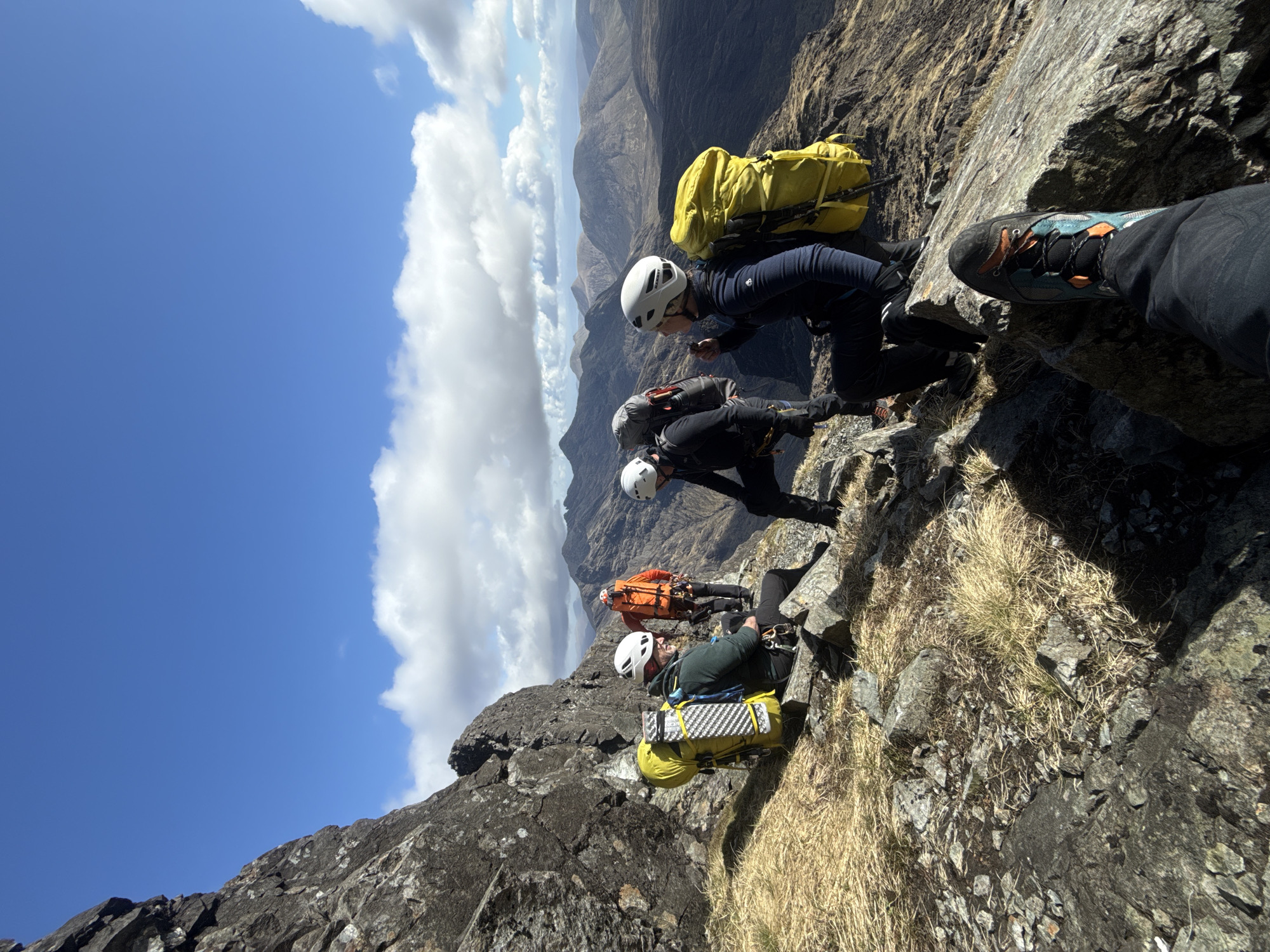 A group of four hikers, all wearing helmets, are traversing a rocky, grassy mountain ridge. The foreground features a close-up of a hiker's boot. The hikers are connected in a line. The sky is blue with scattered white clouds. In the background is a distant mountain range. The location is unknown.